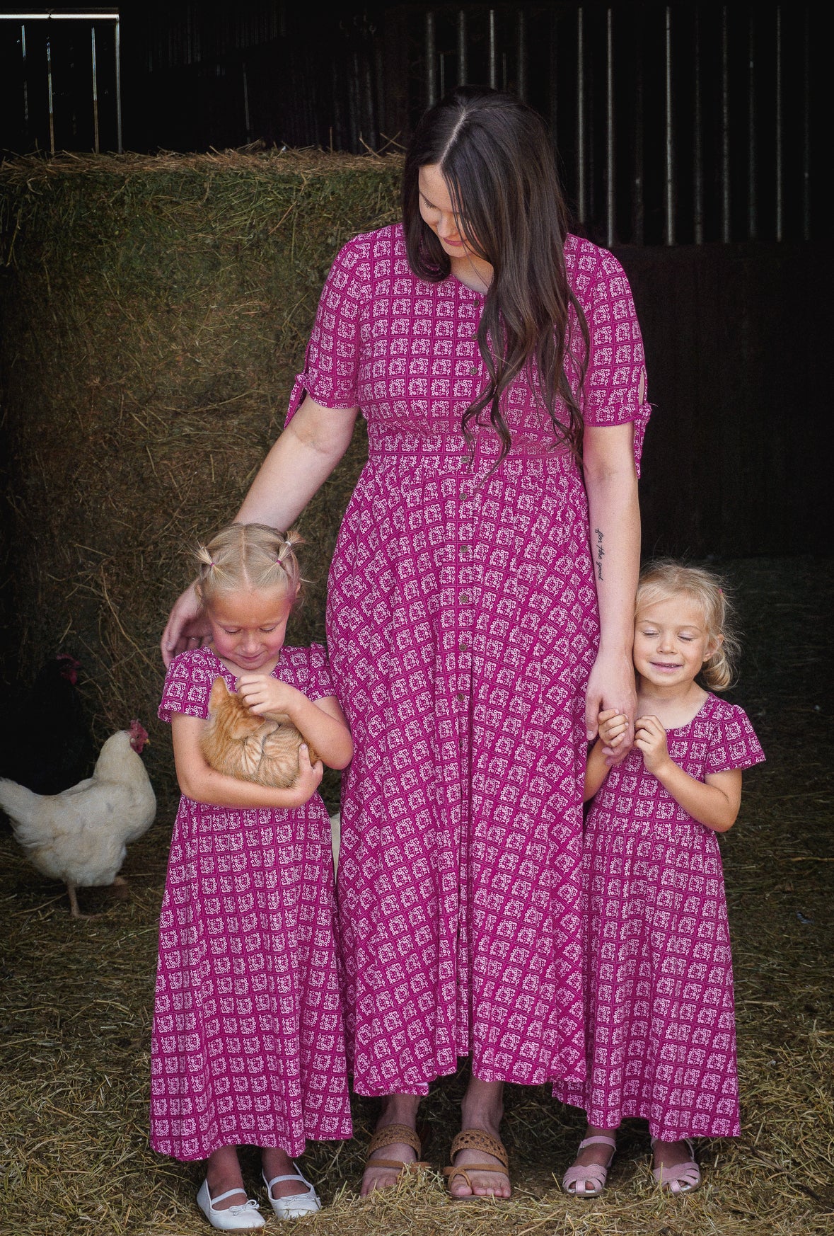 Woman and two young girls in matching pink modest dresses standing on hay with a chicken in the background.