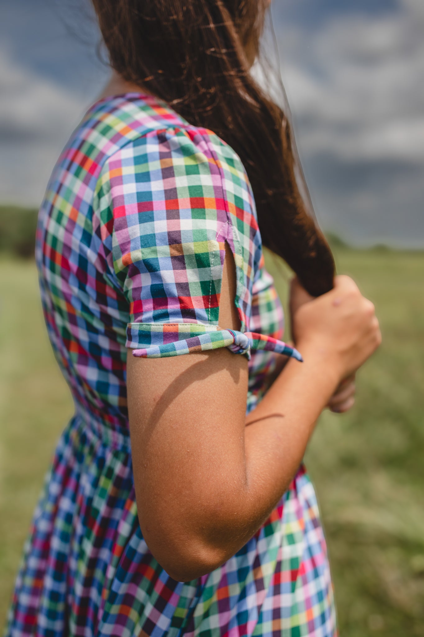 Person wearing a colorful checkered modest nursing dress holding a book in a field with a cloudy sky.