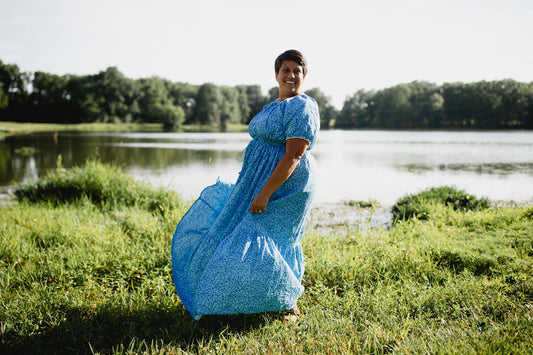 Woman in a blue modest nursing dress standing by a lake with trees in the background
