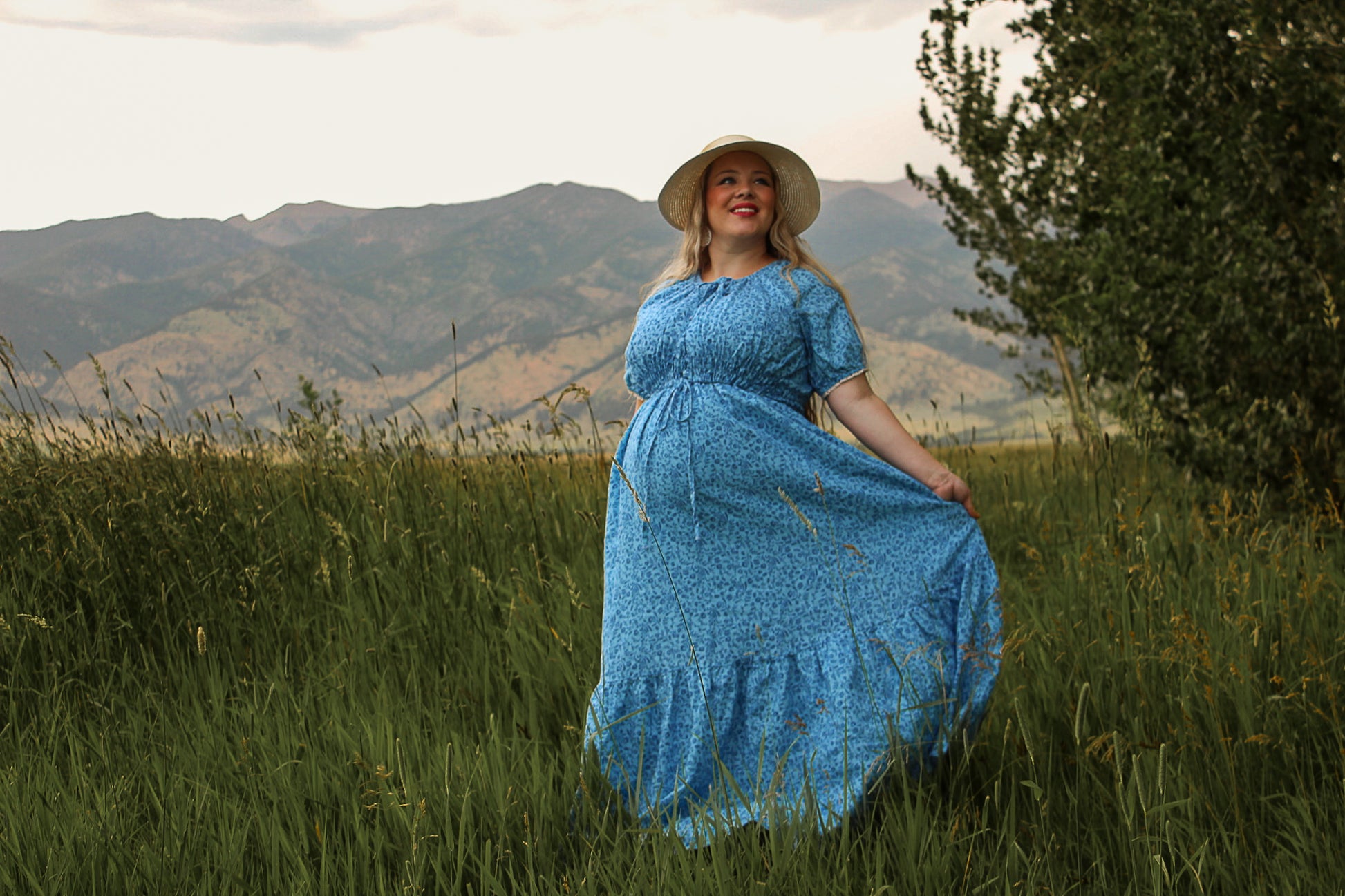 Woman in a blue modest nursing dress and hat standing in a field with mountains in the background