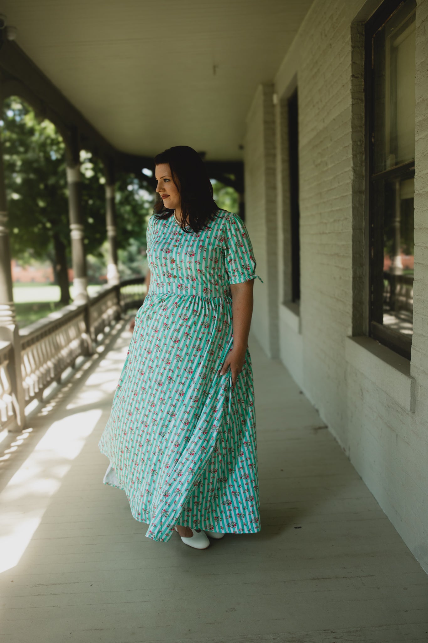 Woman in a green patterned modest nursing dress standing on a porch.