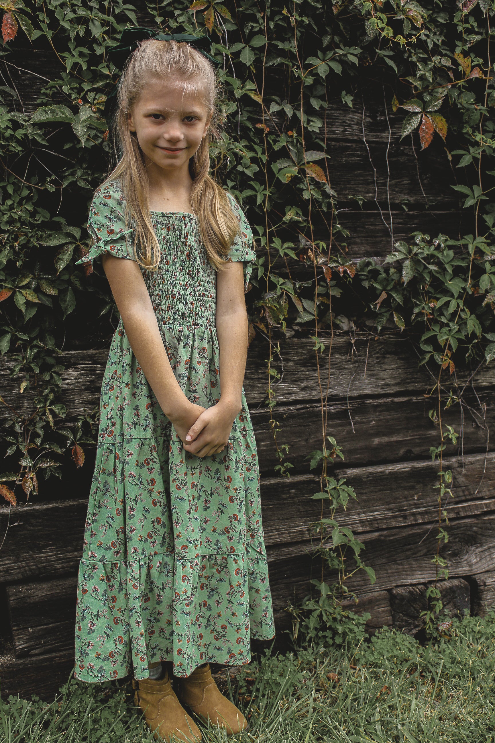 Young girl in a floral dress standing in front of a wooden wall with greenery.