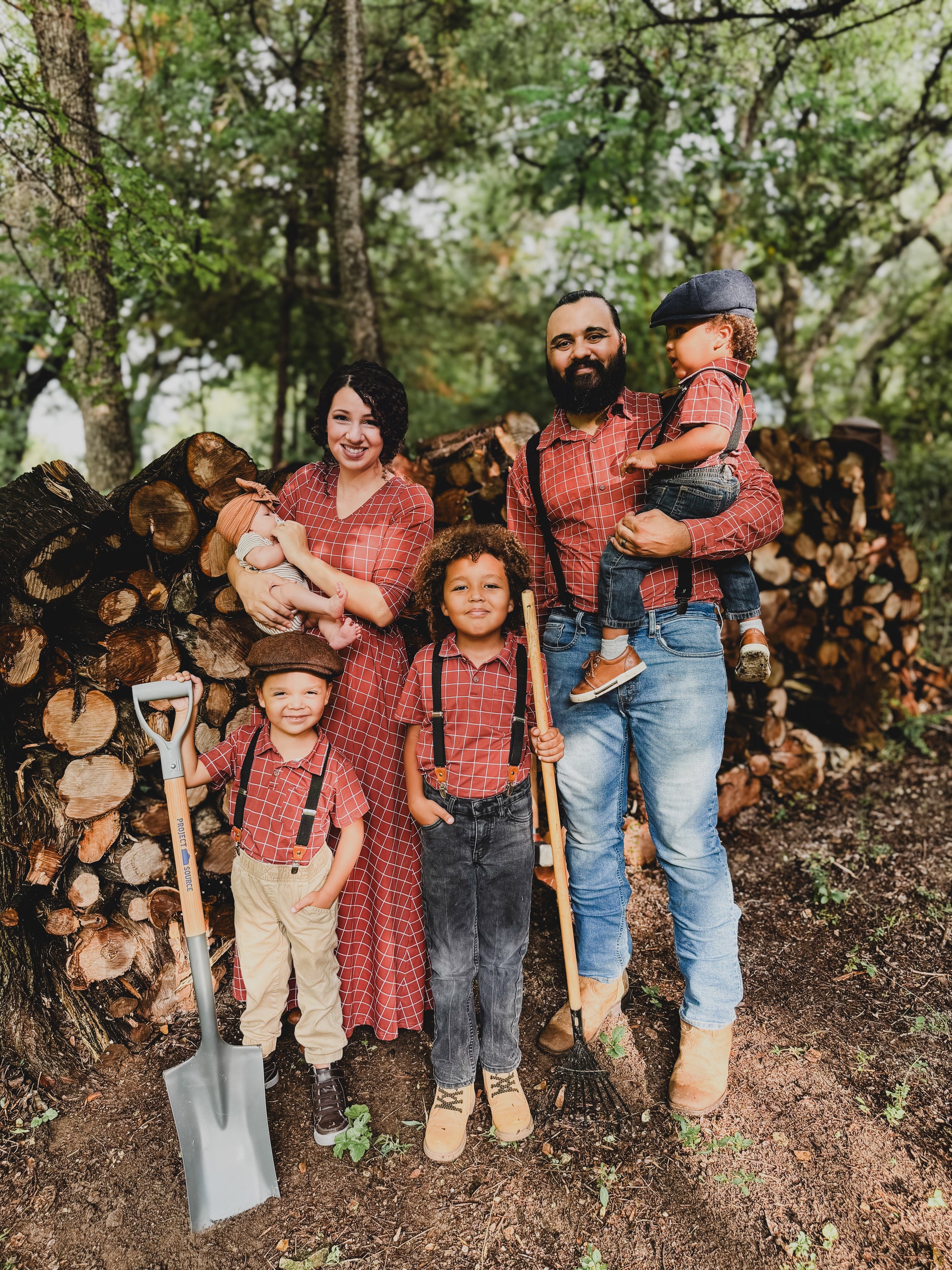 Family of five wearing modest copper windowpane plaid maxi dresses posing together outdoors by rustic wood pile, featuring mother in nursing-friendly dress with daughters in matching dresses, showcasing the comfortable cotton blend fabric and coordinated family style perfect for outdoor photoshoots