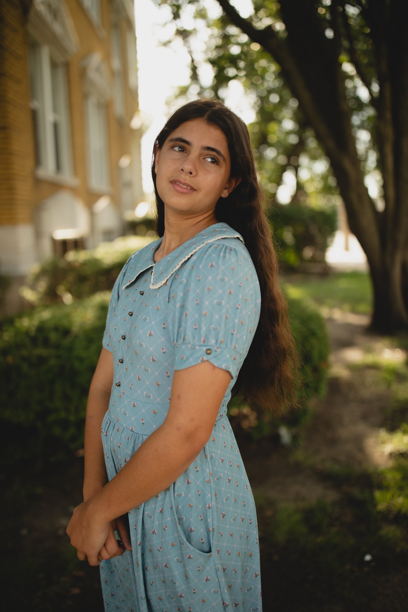 Woman in a blue modest dress standing outdoors with greenery and a building in the background