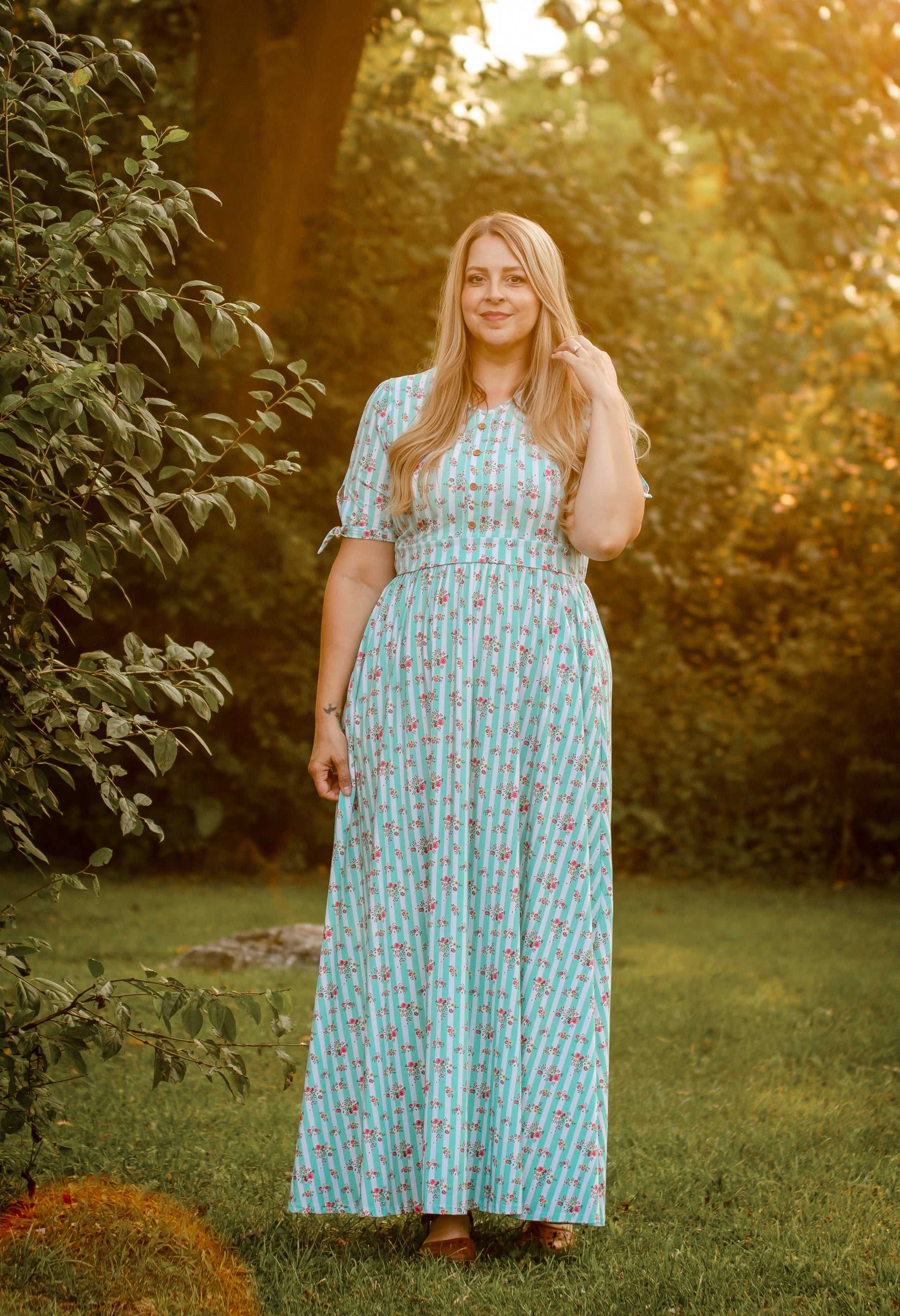 Woman in a patterned modest dress standing in a garden with trees and grass.