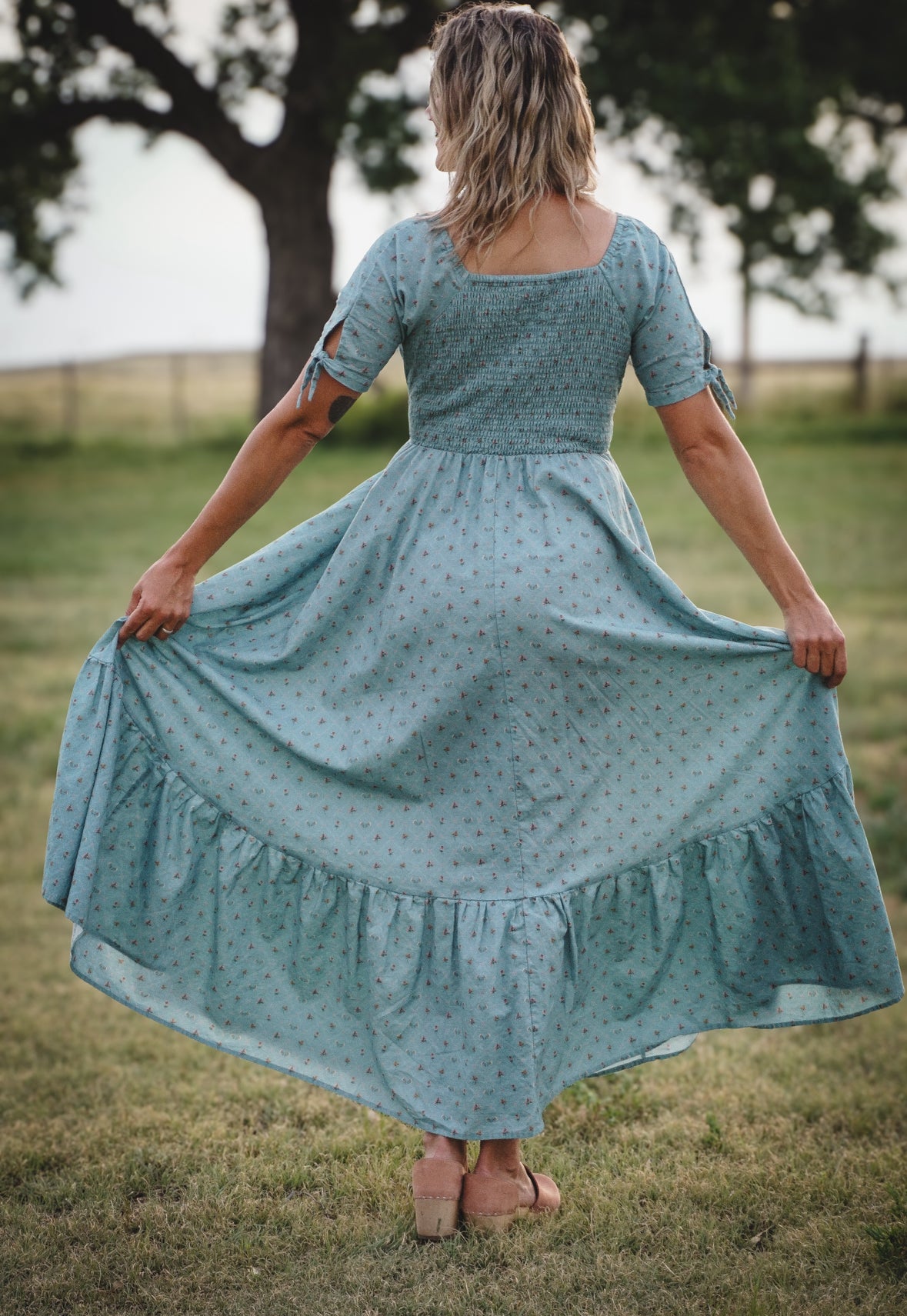 Woman in a blue modest nursing dress standing in a grassy field with trees in the background
