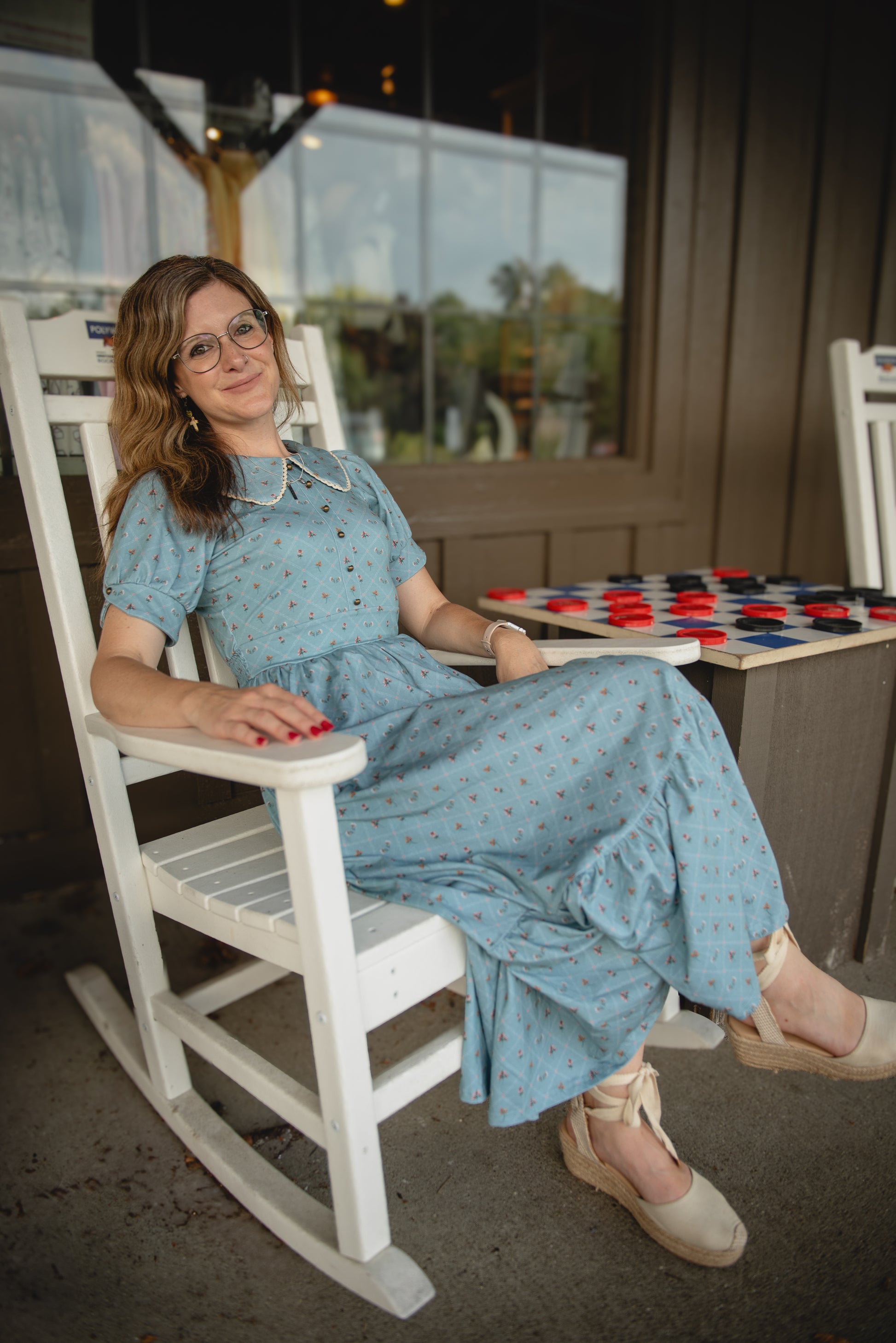 Woman in a blue modest nursing dress sitting on a white rocking chair in a casual setting.