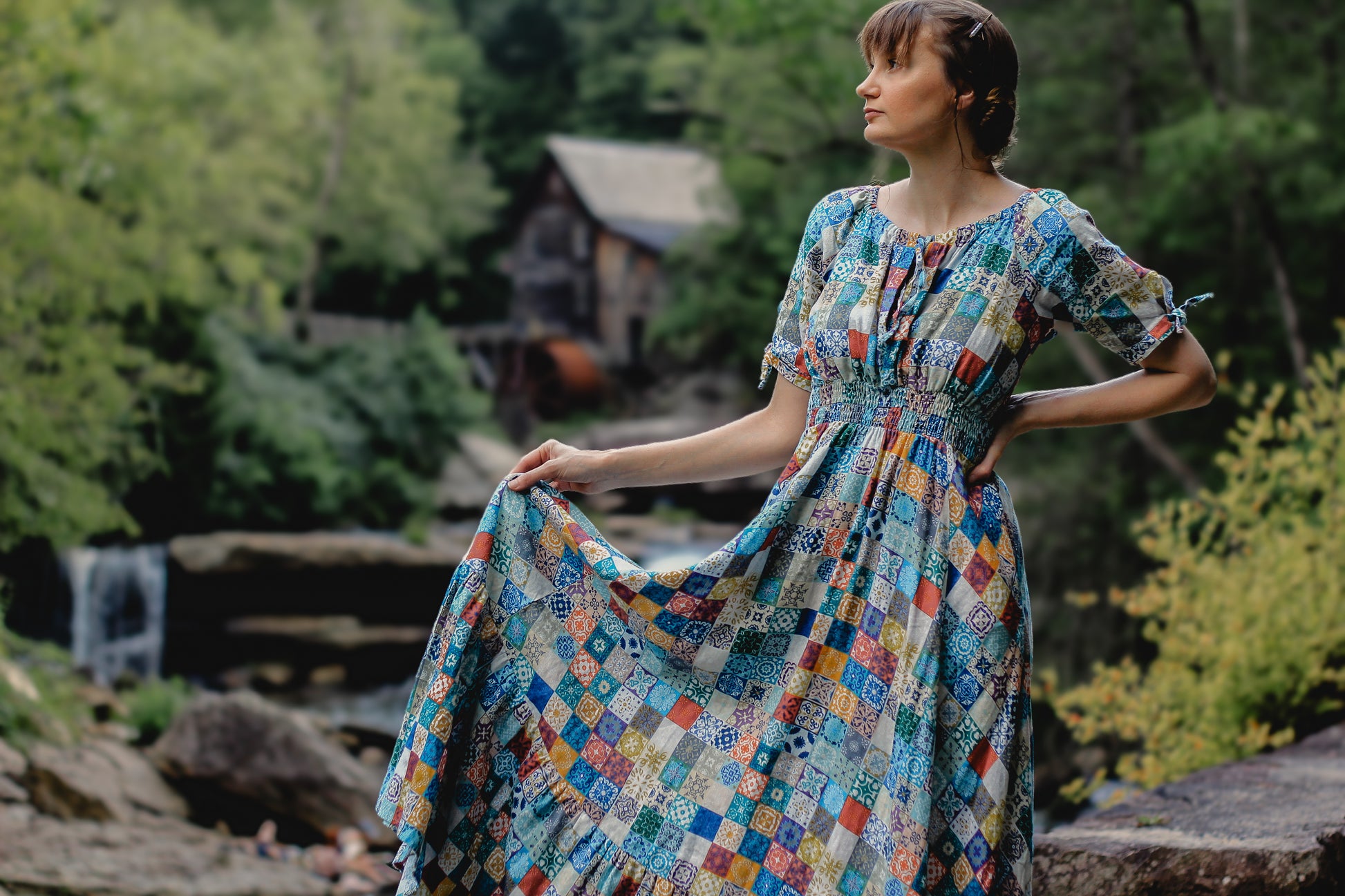 Woman in a colorful modest nursing dress standing in a natural setting with trees and a building in the background.