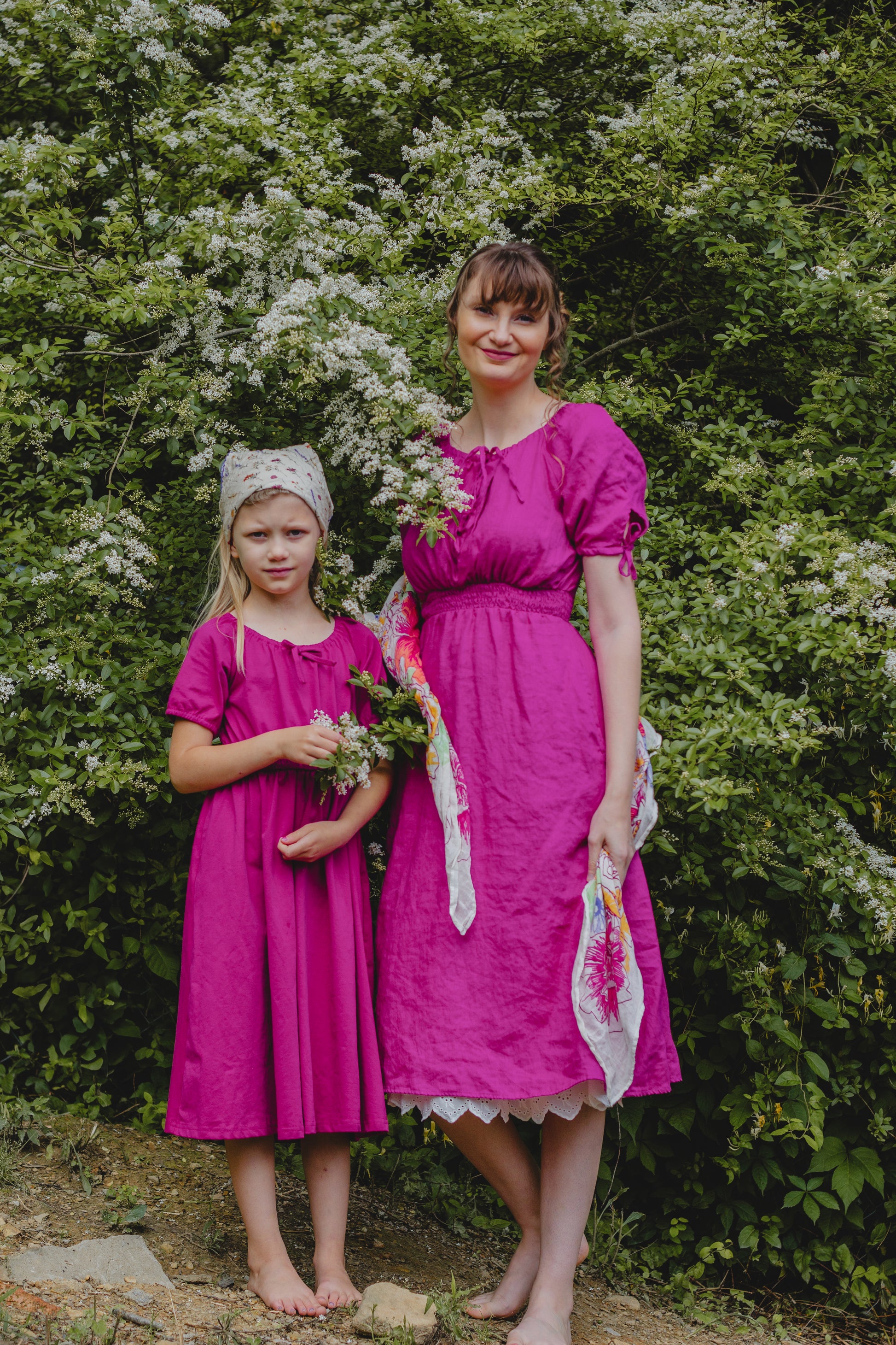 Mother and daughter in pink modest dresses standing in a garden with greenery and flowers.