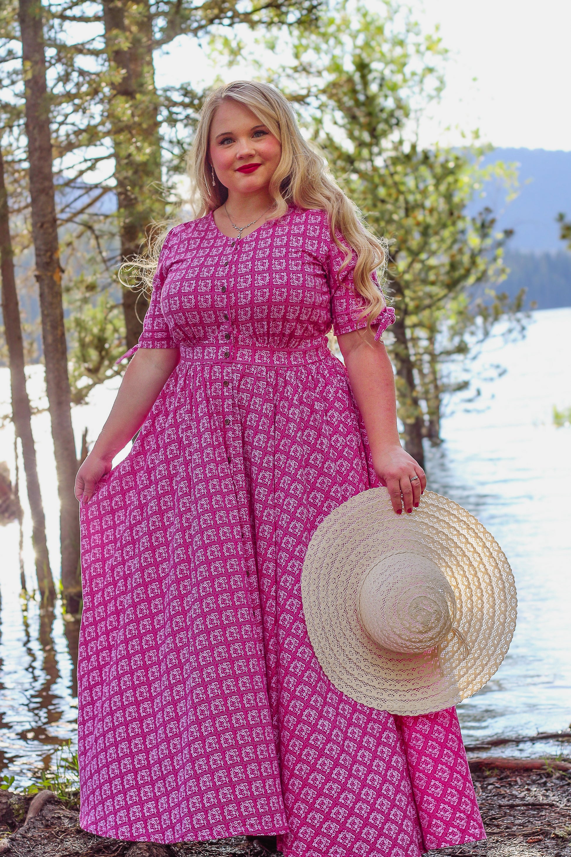 Woman in a pink modest nursing dress holding a straw hat by a lake with trees in the background