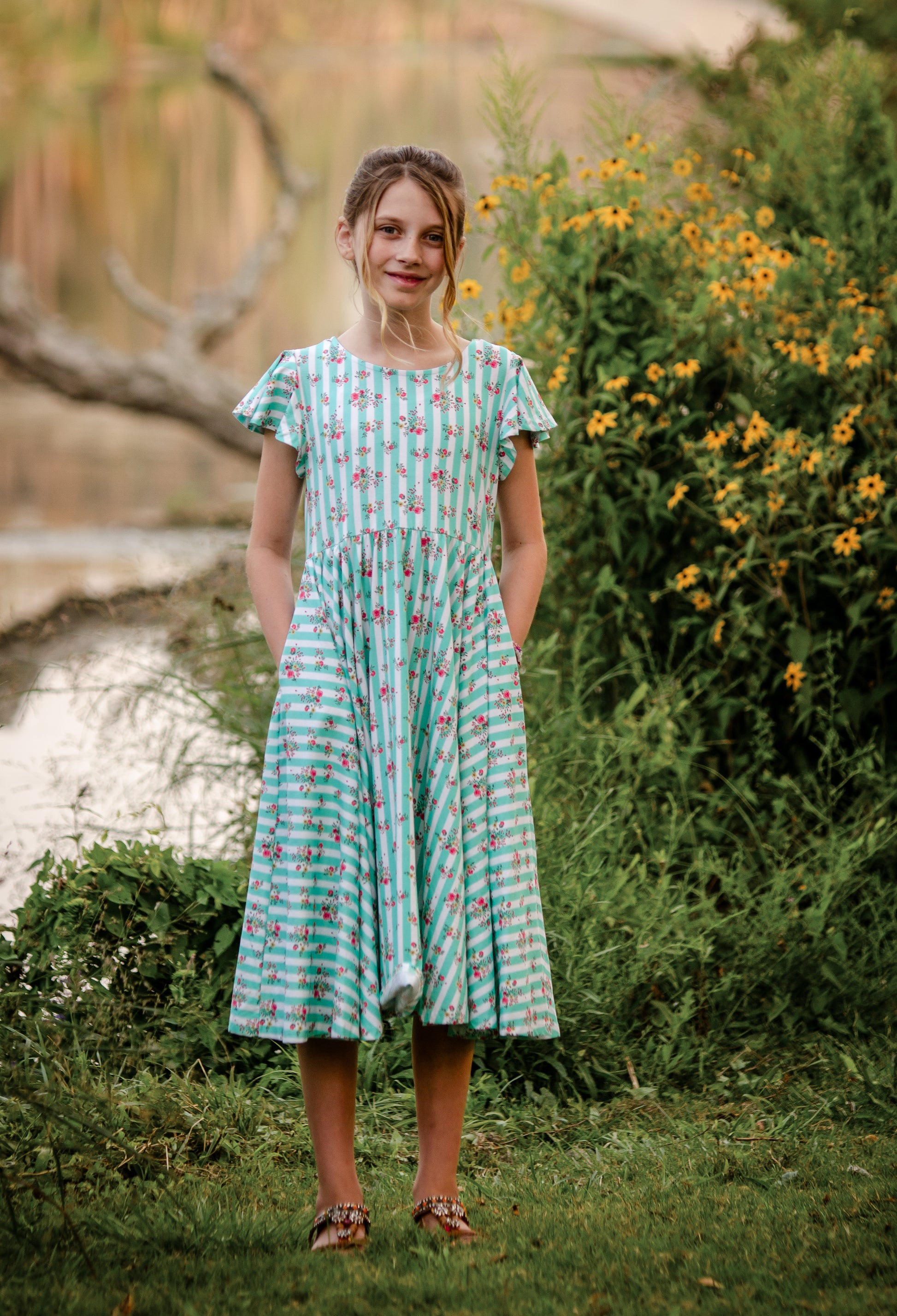 Young girl in a green modest dress with floral pattern standing outdoors near a body of water and flowers.