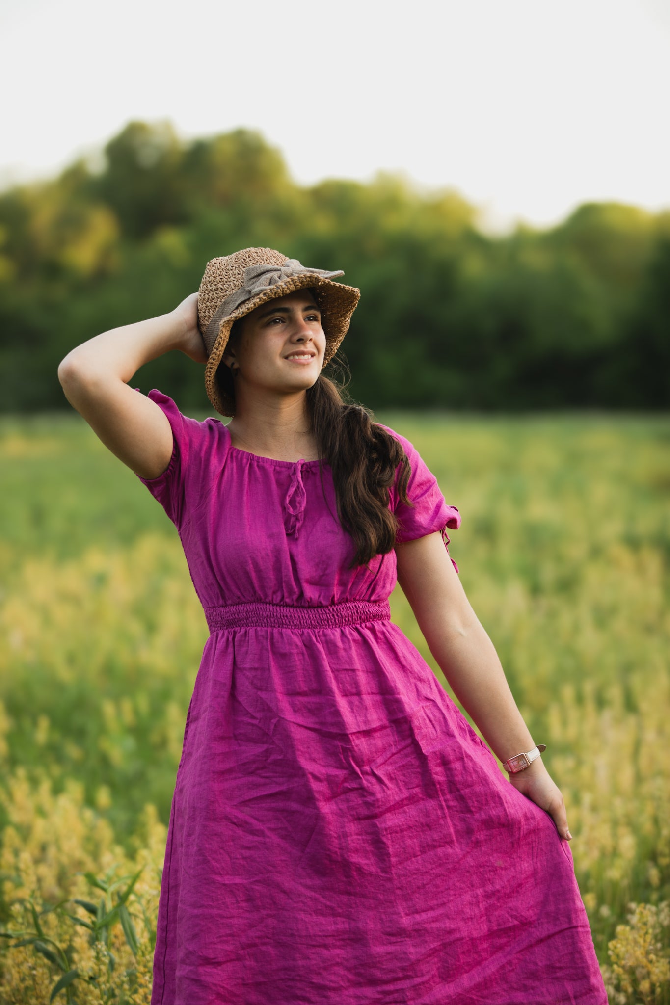 Woman in a purple modest nursing dress and straw hat standing in a field with trees in the background