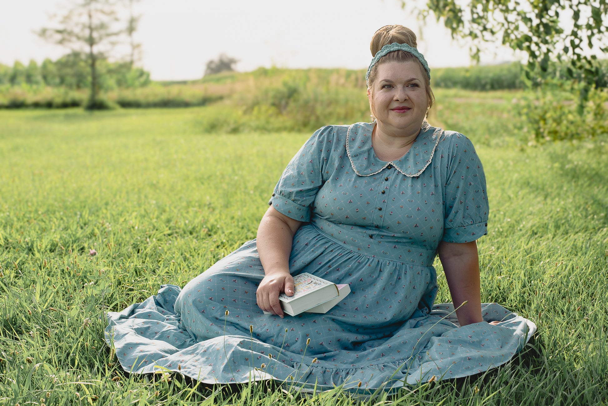 Woman in a blue modest nursing dress sitting on grass holding a book