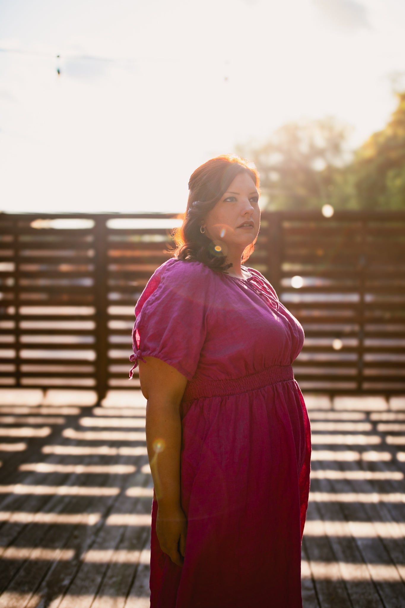 Woman in a pink modest nursing dress standing on a wooden deck with sunlight filtering through.
