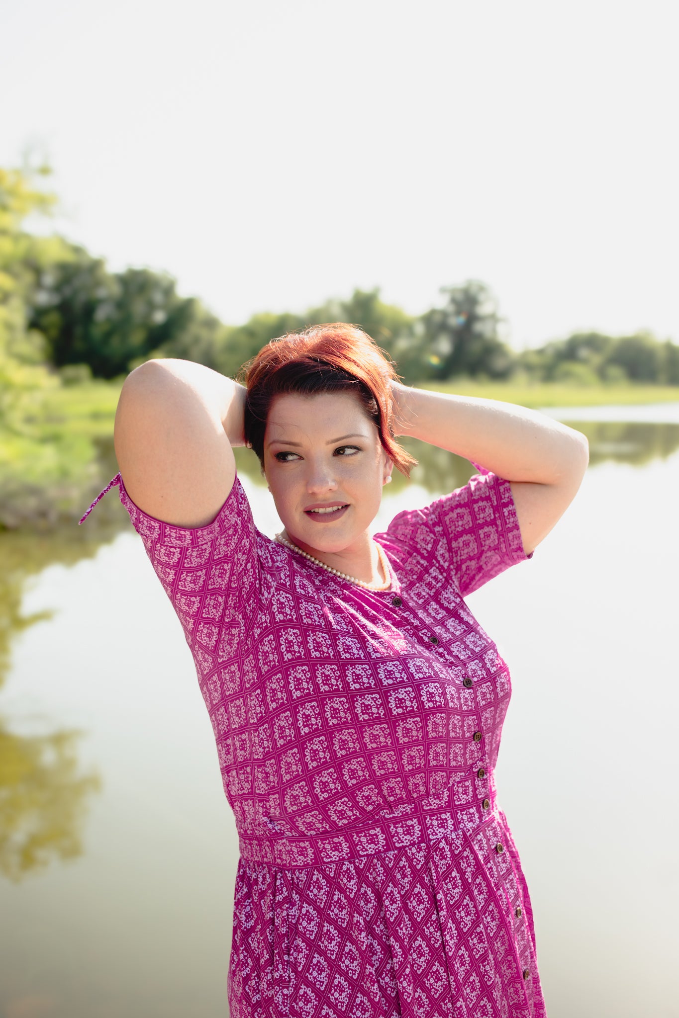Woman in a pink patterned modest nursing dress standing by a body of water with trees in the background