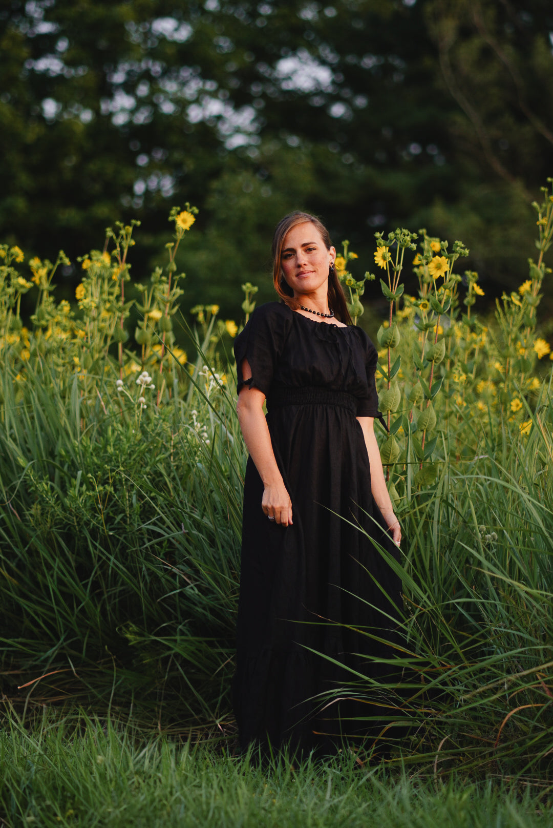 Woman in a black modest nursing dress standing in a field of tall grass and wildflowers.