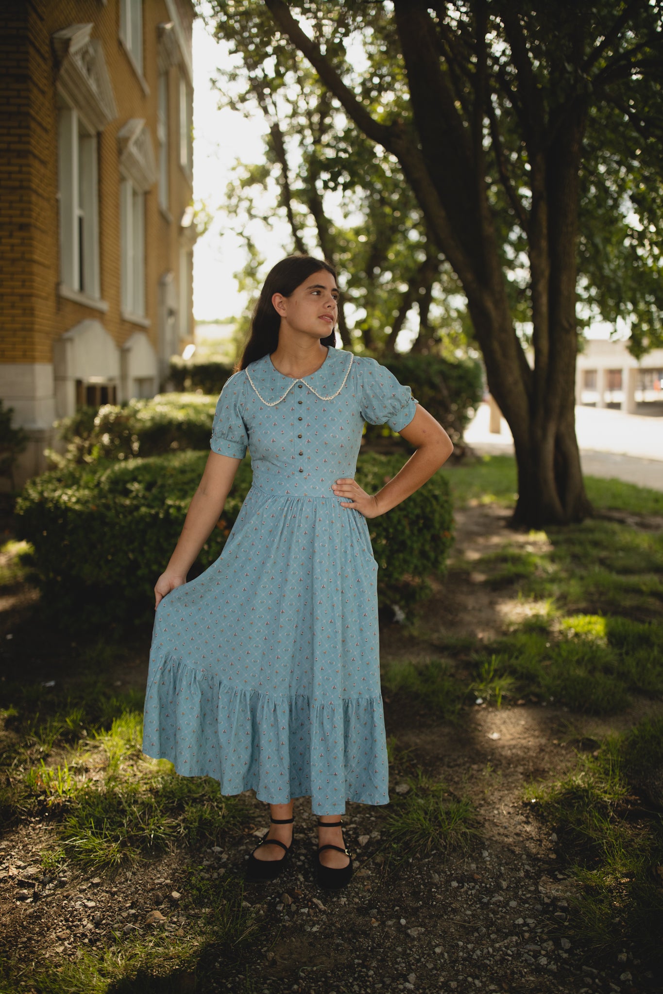 Woman in a blue modest nursing dress standing outdoors near a building and tree
