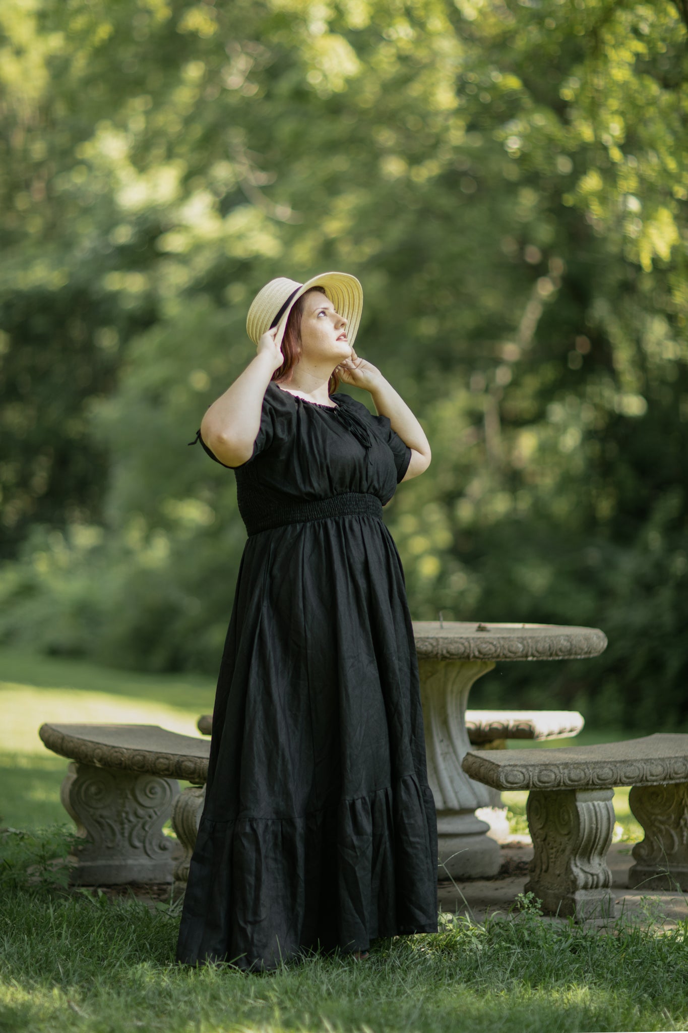 Woman in a black modest nursing dress and straw hat standing in a park with stone benches and greenery.