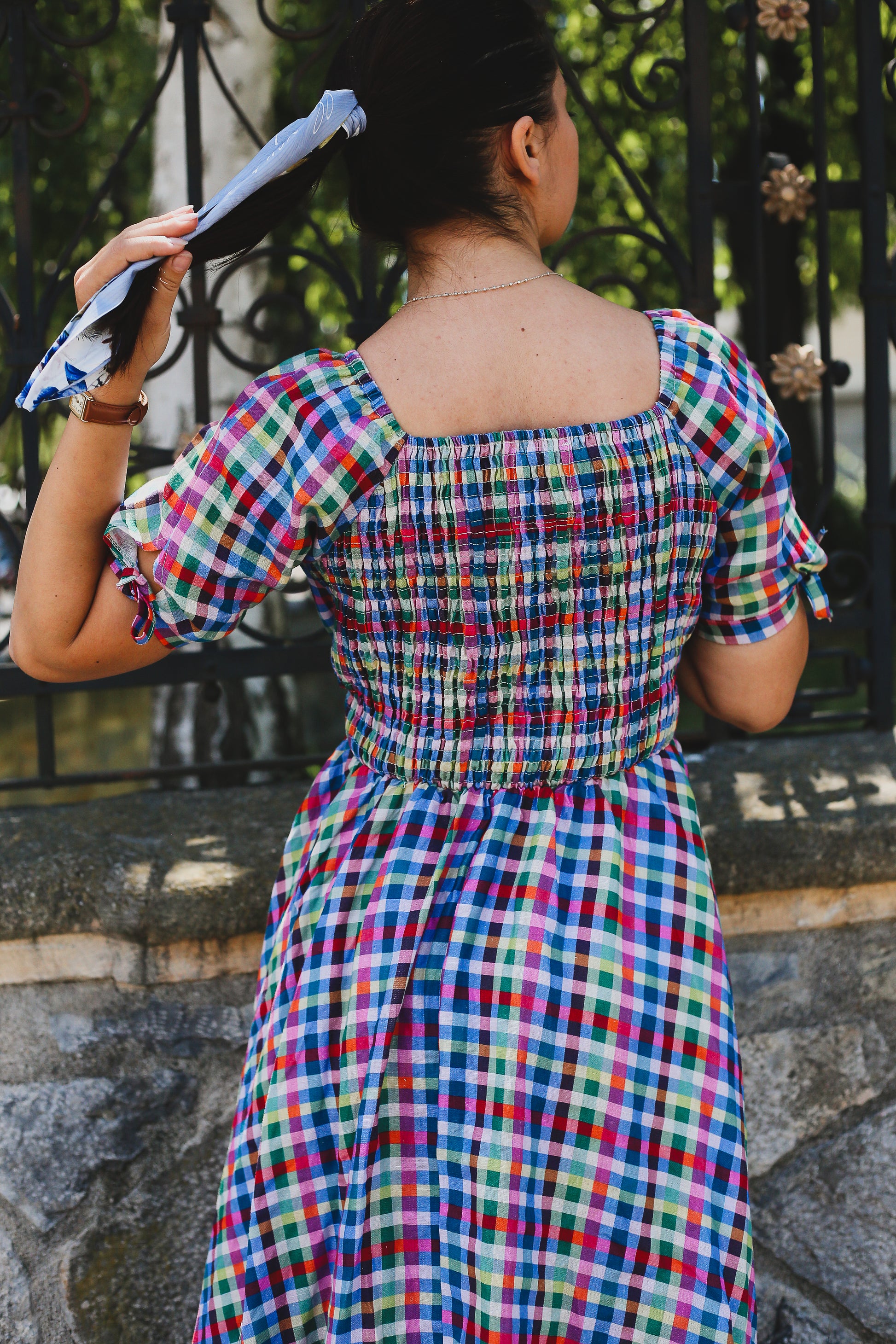 Woman wearing a colorful checkered modest nursing dress standing against a stone wall with a metal gate.