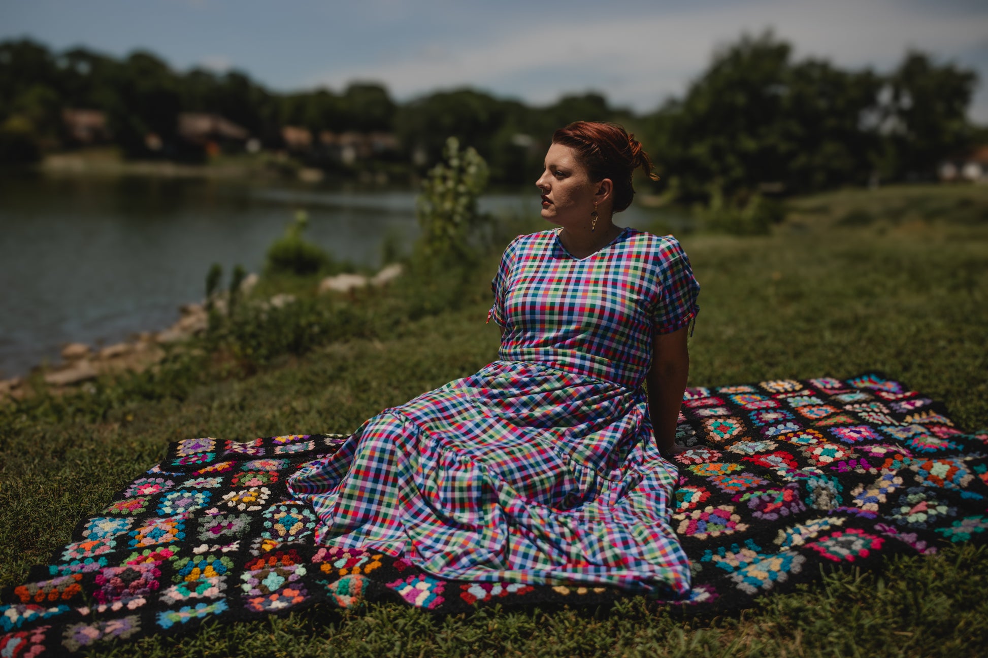 Woman in a colorful modest nursing dress sitting on a large crocheted blanket by a lake.