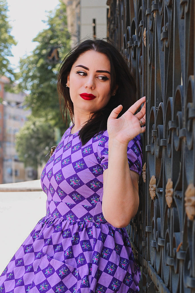 Woman in a purple checkered modest dress leaning against a fence outdoors.
