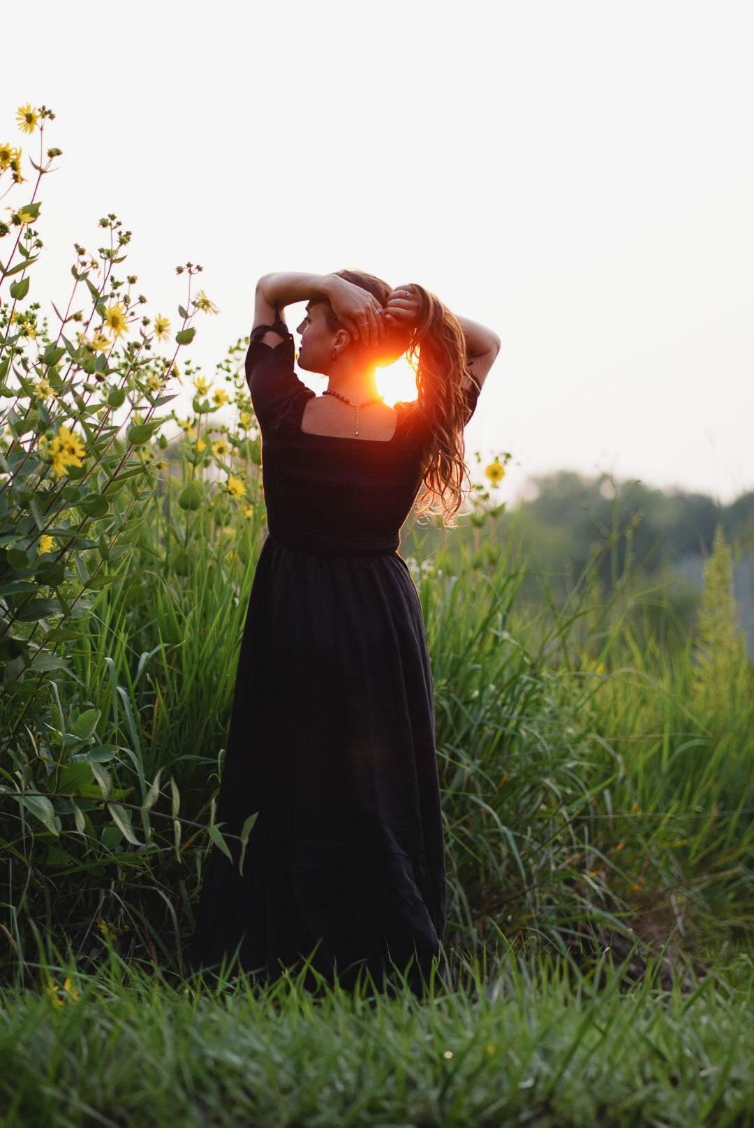 Person in a black modest nursing dress standing in a field with the sun setting behind them