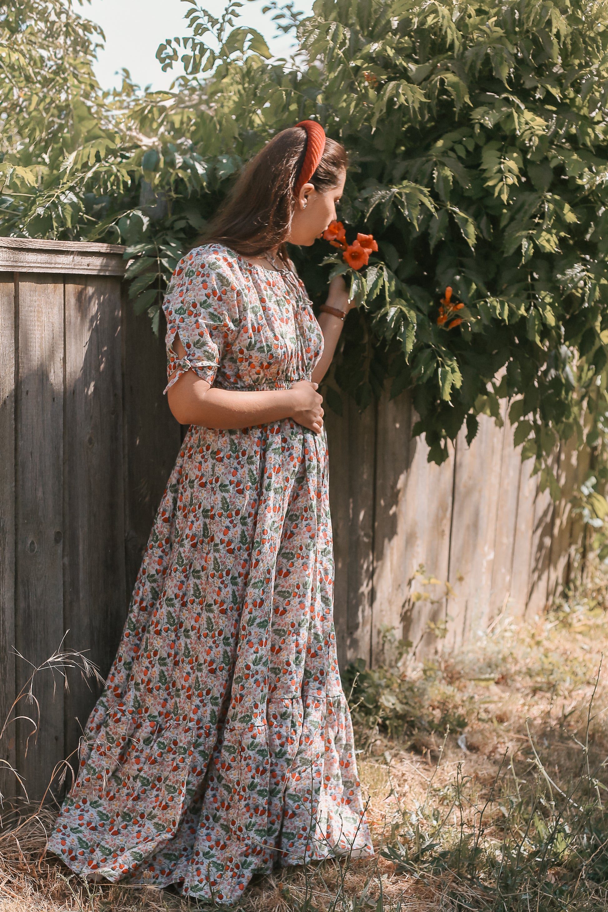 Woman in a floral modest nursing dress standing outdoors near a wooden fence and greenery.