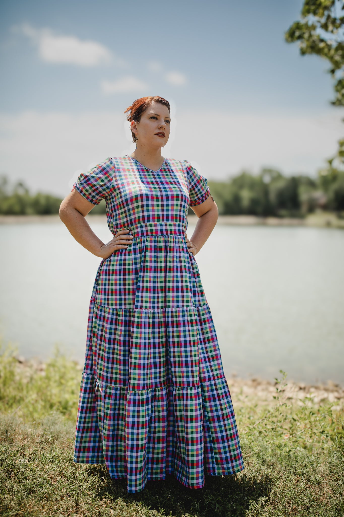 Woman in a plaid modest nursing dress standing by a body of water with trees in the background