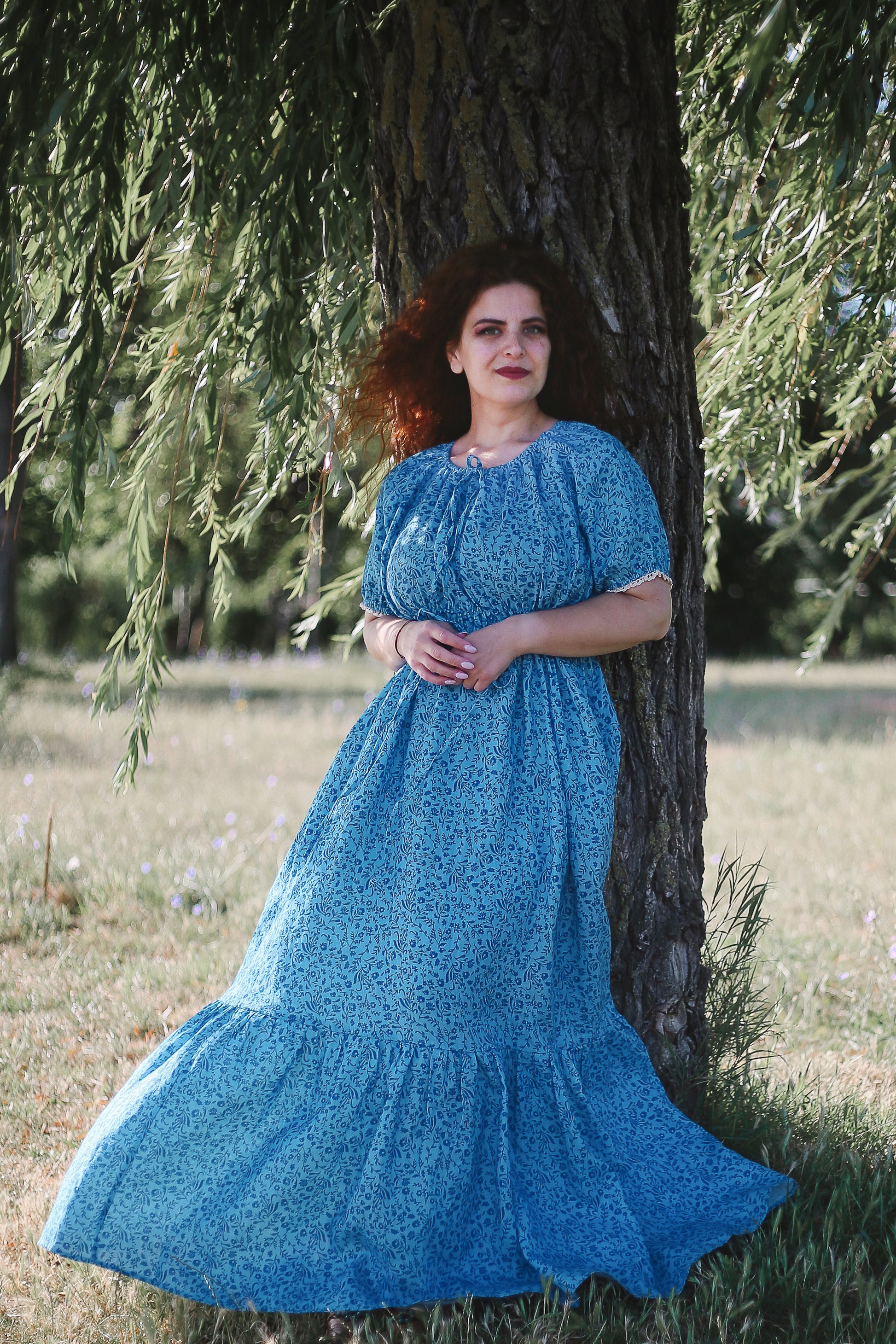 Woman in a blue modest nursing dress standing next to a tree in a natural setting