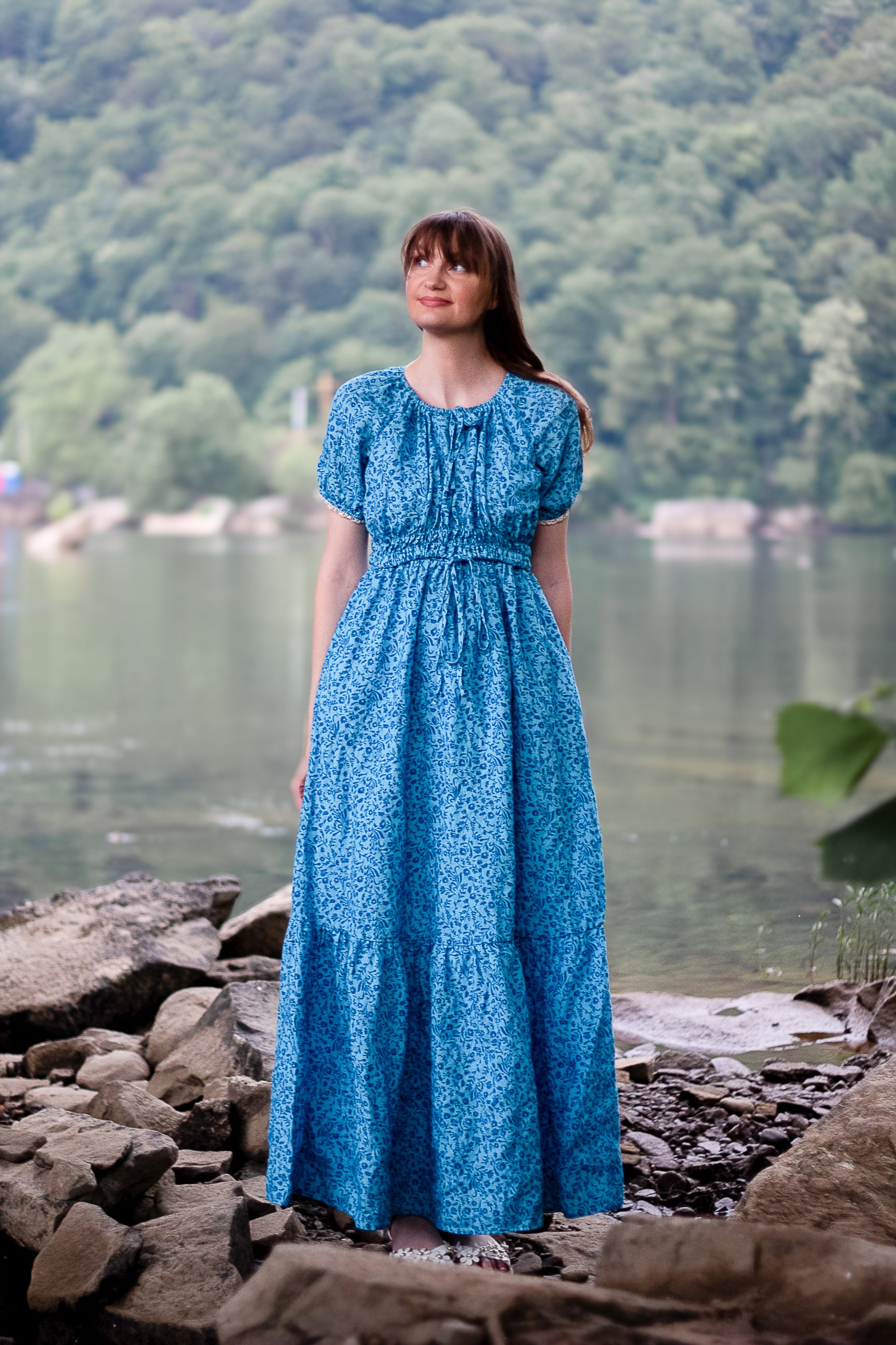 Woman in a blue modest nursing dress standing by a lake with mountains in the background