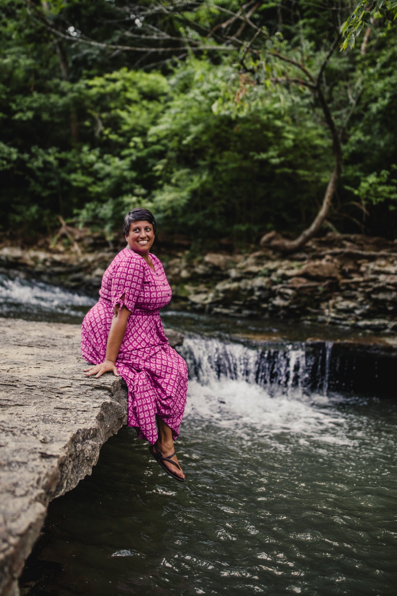 Woman in a pink modest nursing dress sitting on a rock by a small waterfall surrounded by greenery.