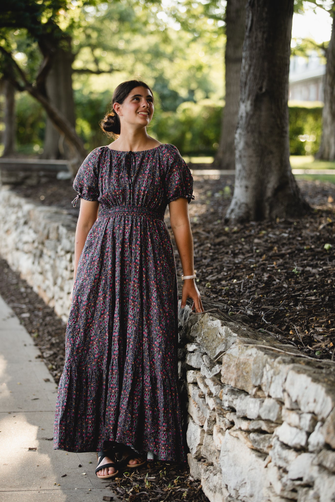 Woman in a long, patterned modest nursing dress standing by a stone wall in a garden.