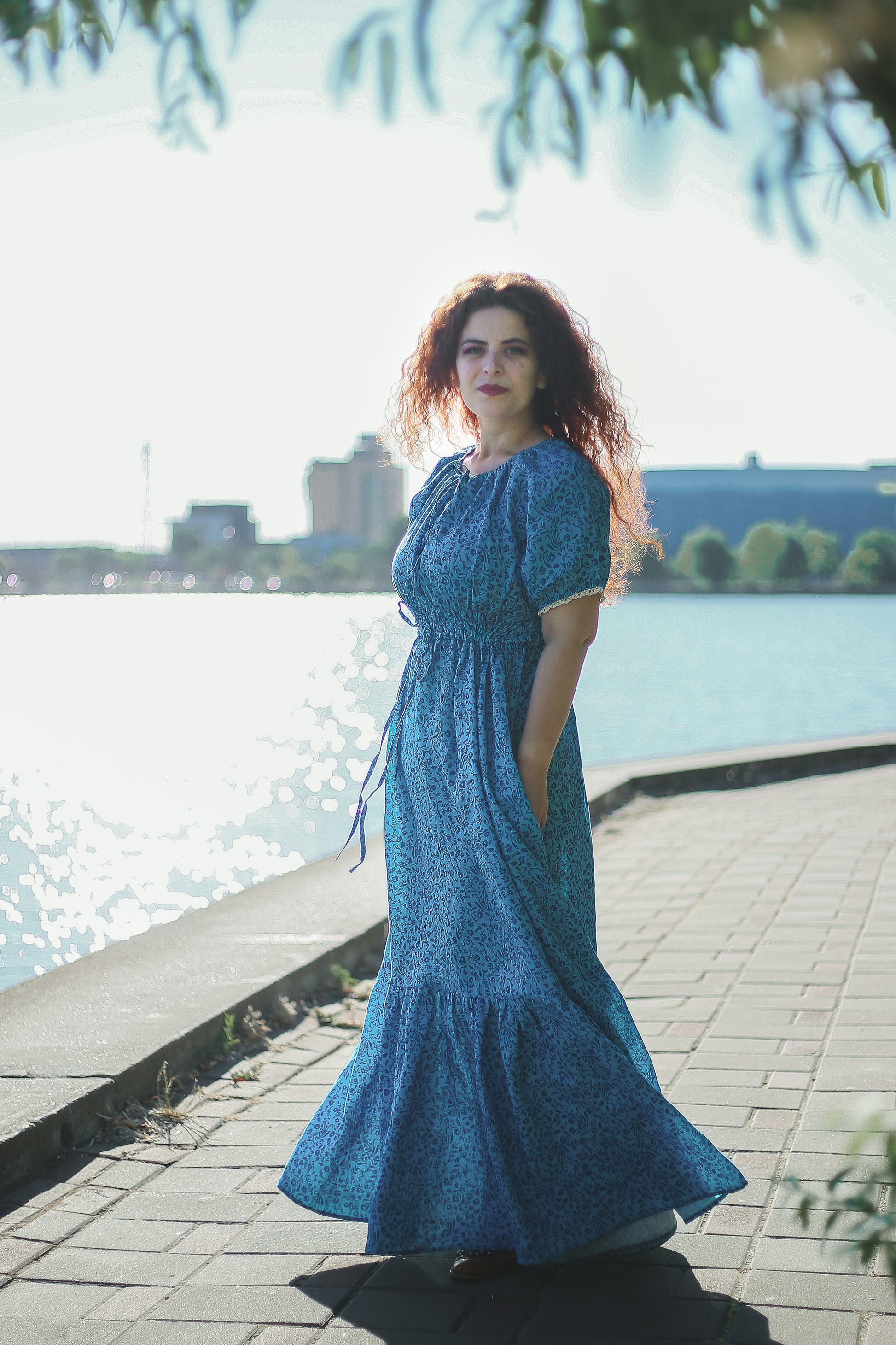 Woman in a blue modest nursing dress standing by a waterfront with buildings in the background