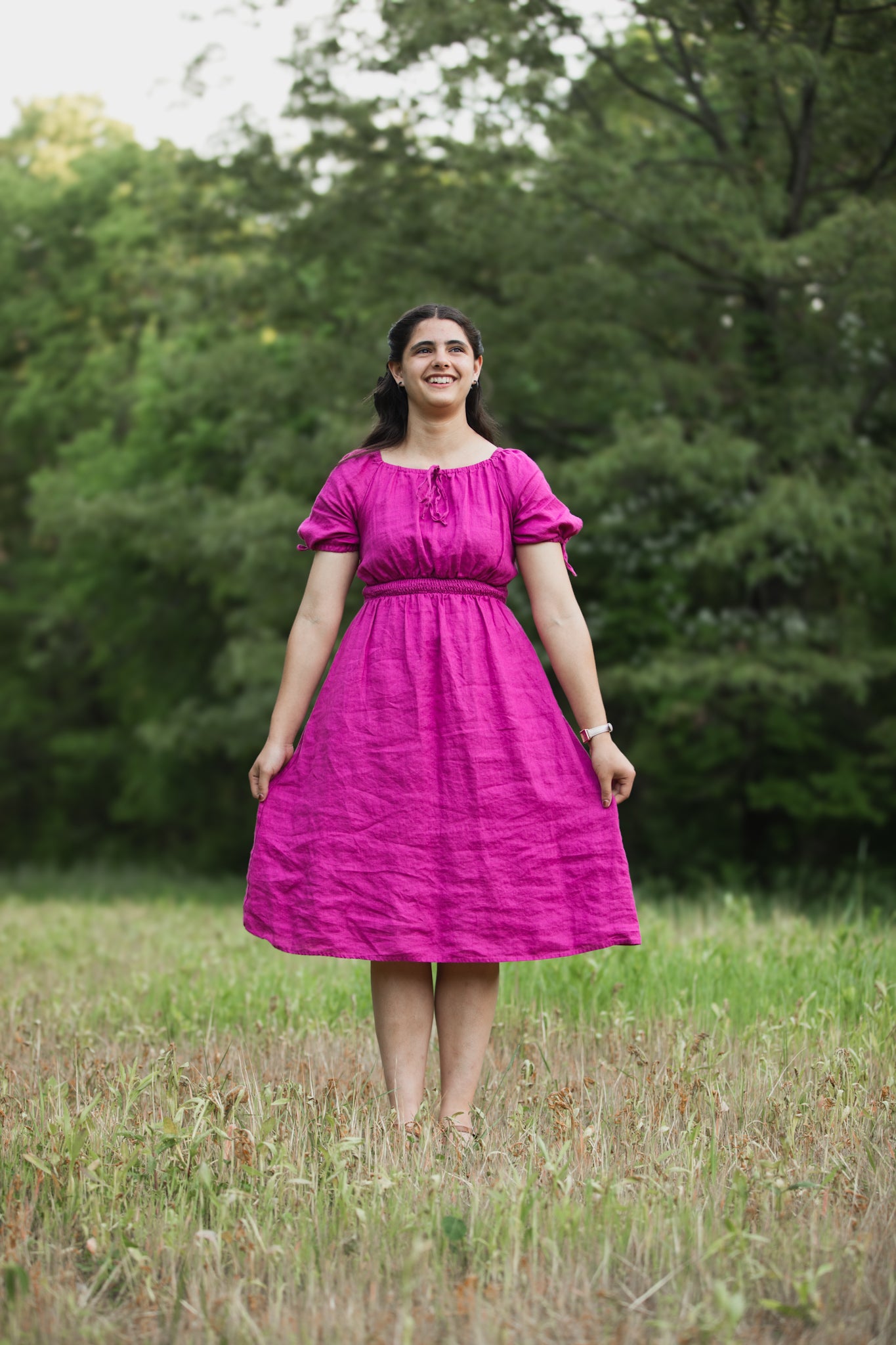 Woman in a pink modest nursing dress standing in a grassy field with trees in the background