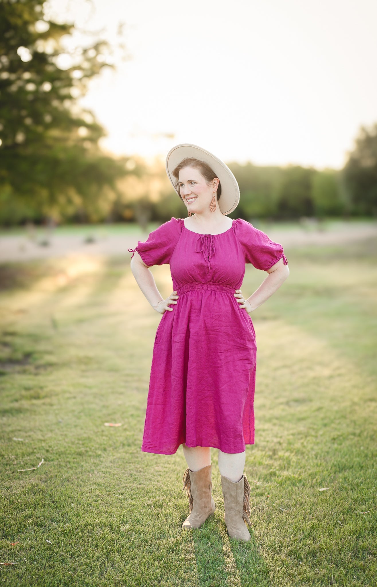 Woman in a pink modest nursing dress and hat standing in a grassy field.