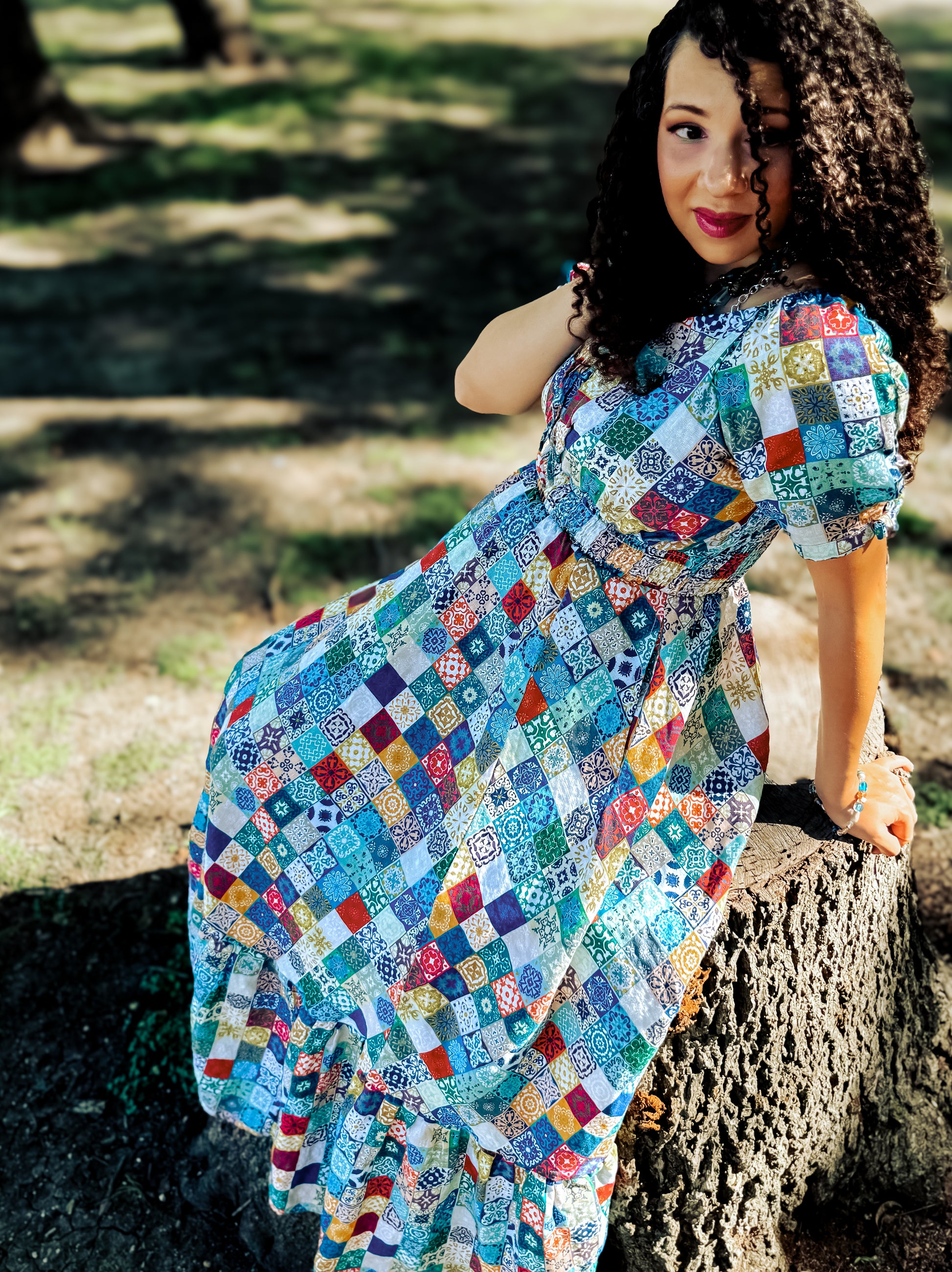 Woman wearing a colorful patchwork modest nursing dress sitting on a tree stump outdoors.