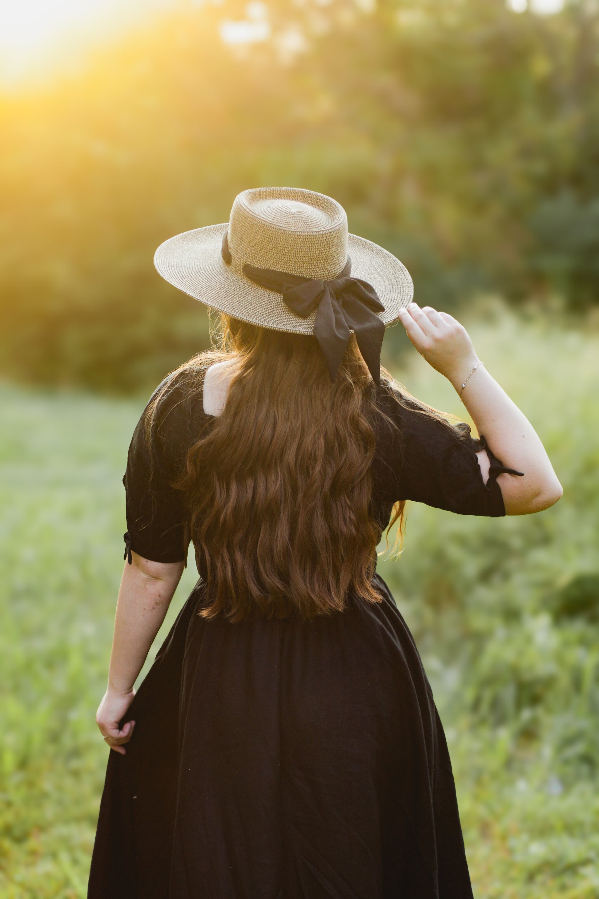 Woman in a black modest nursing dress and straw hat standing in a field with sunlight filtering through.