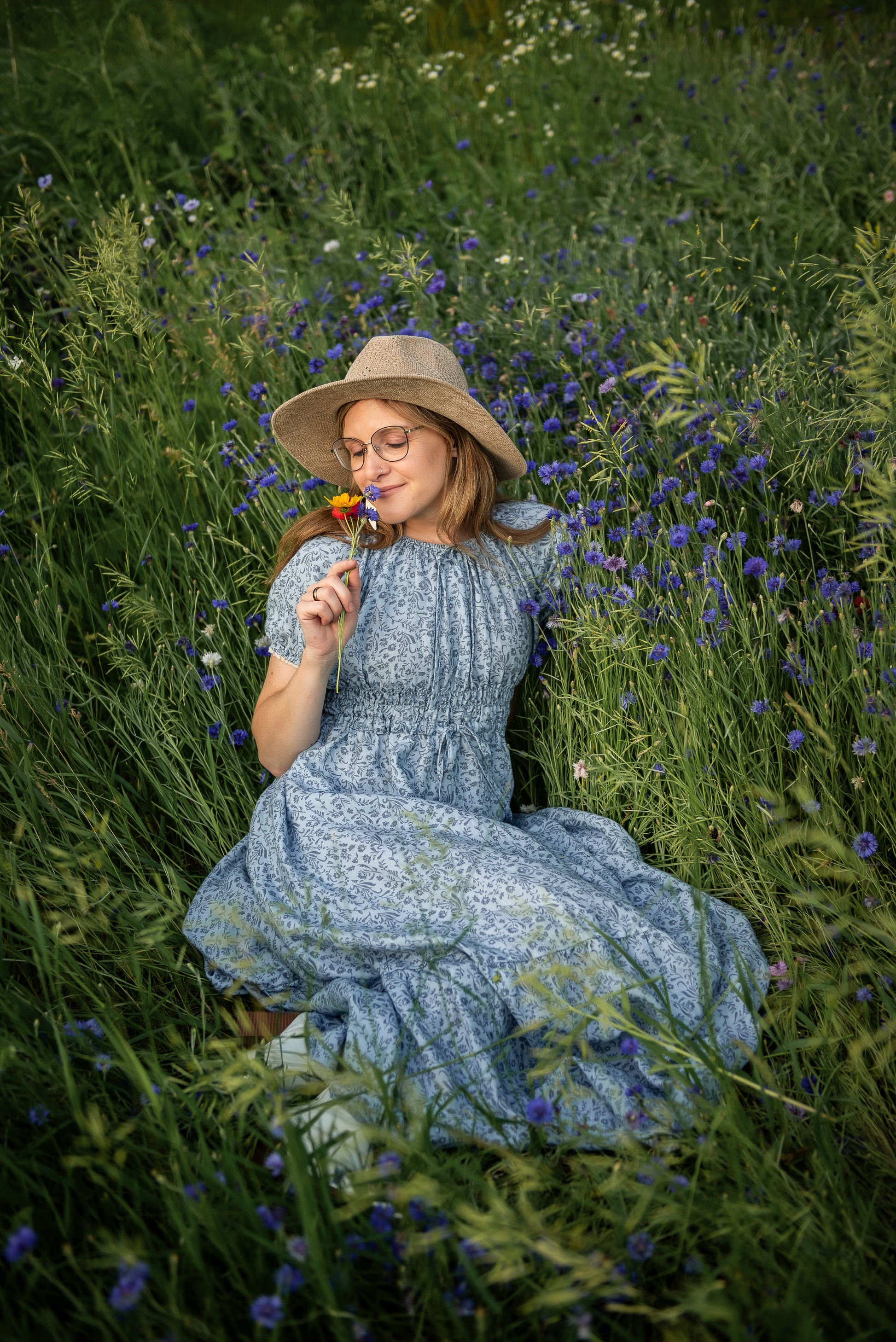 Woman in a blue modest blue nursing dress and hat sitting in a field of wildflowers