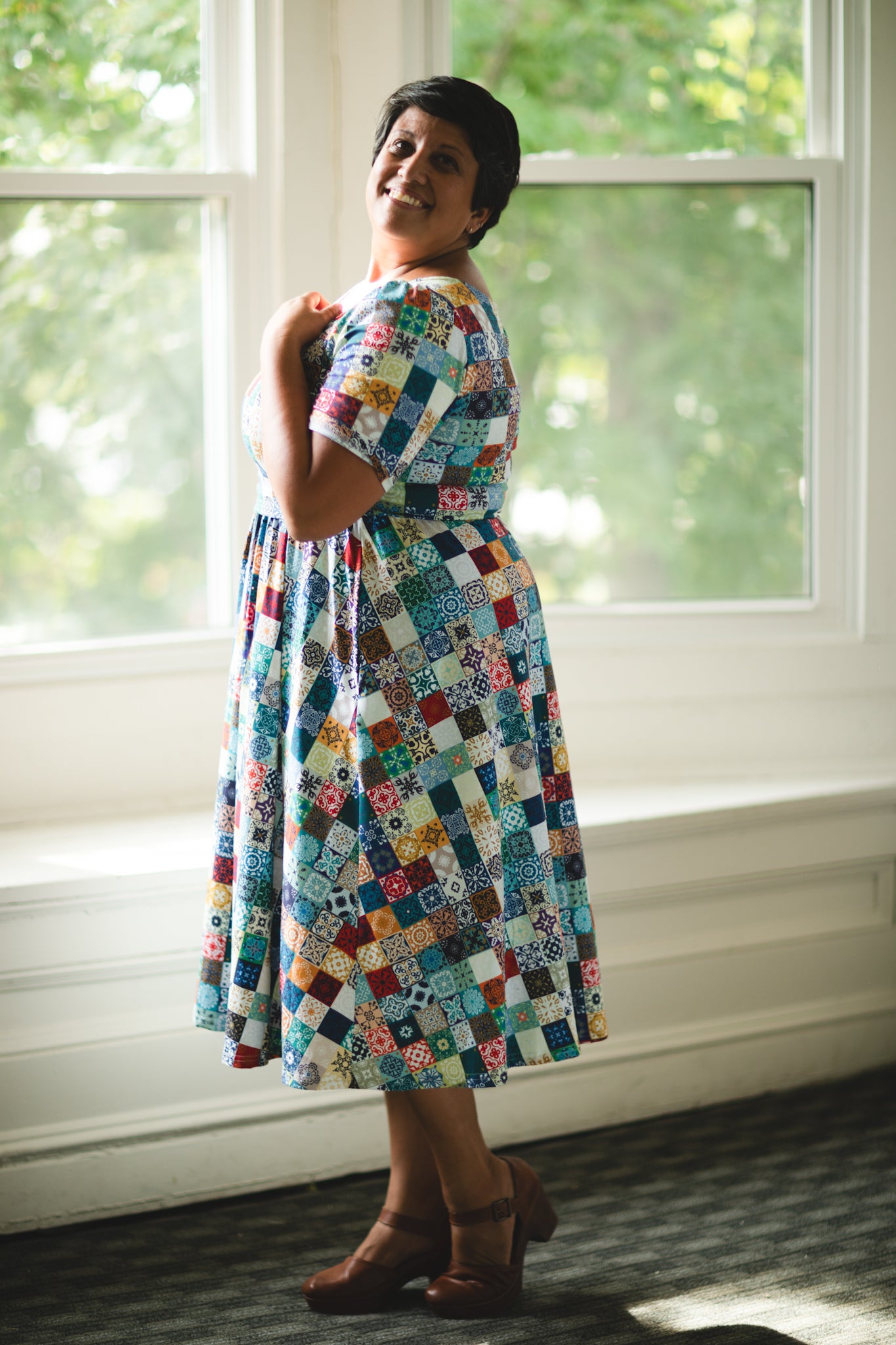 Woman wearing a colorful patchwork modest nursing dress standing in a room with large windows.