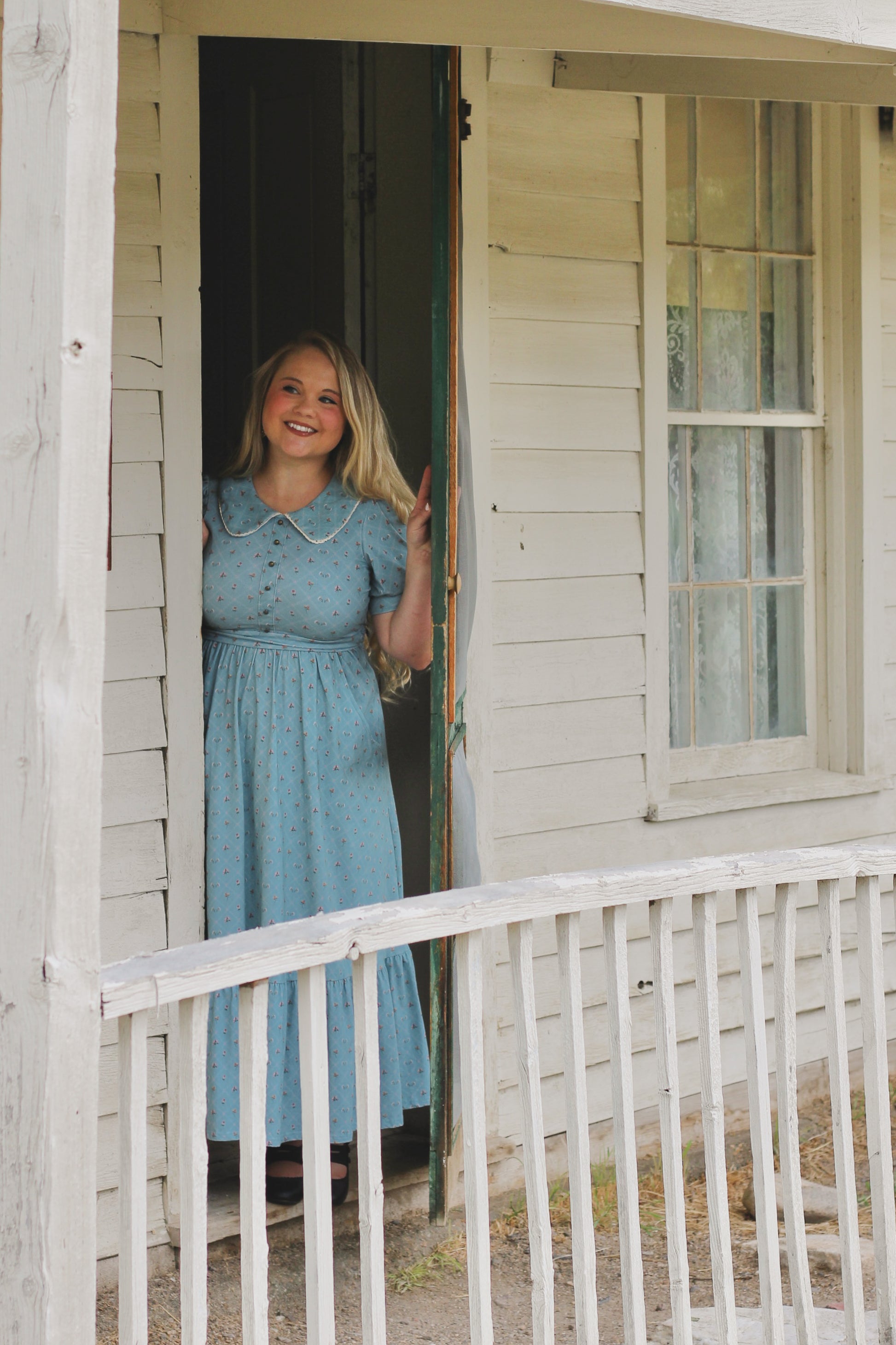 Woman in a blue modest nursing dress standing on a porch of a house