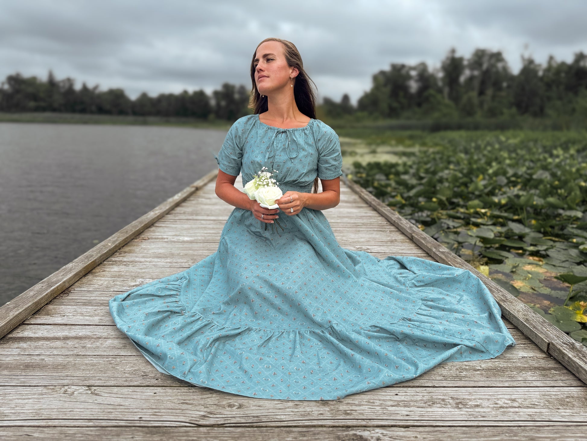 Woman in a blue modest nursing dress standing on a wooden dock by a lake