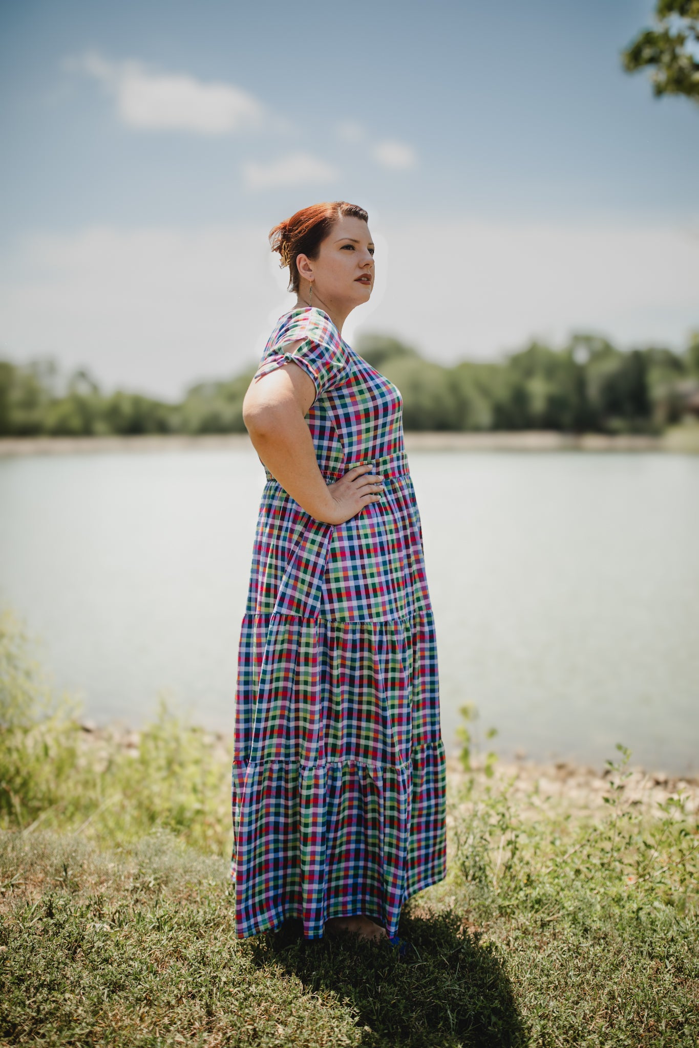 Woman in a plaid modest nursing dress standing by a body of water with trees in the background