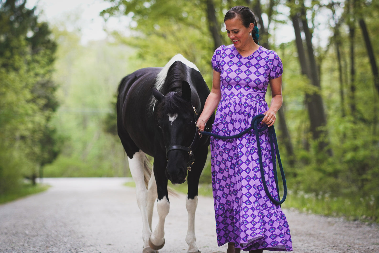 Woman in a purple modest nursing dress walking a horse on a path in the woods.