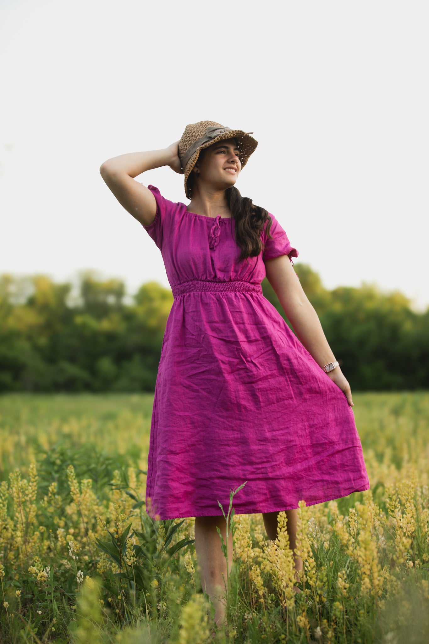 Woman in a pink modest nursing dress and hat standing in a field with greenery and yellow flowers in the background
