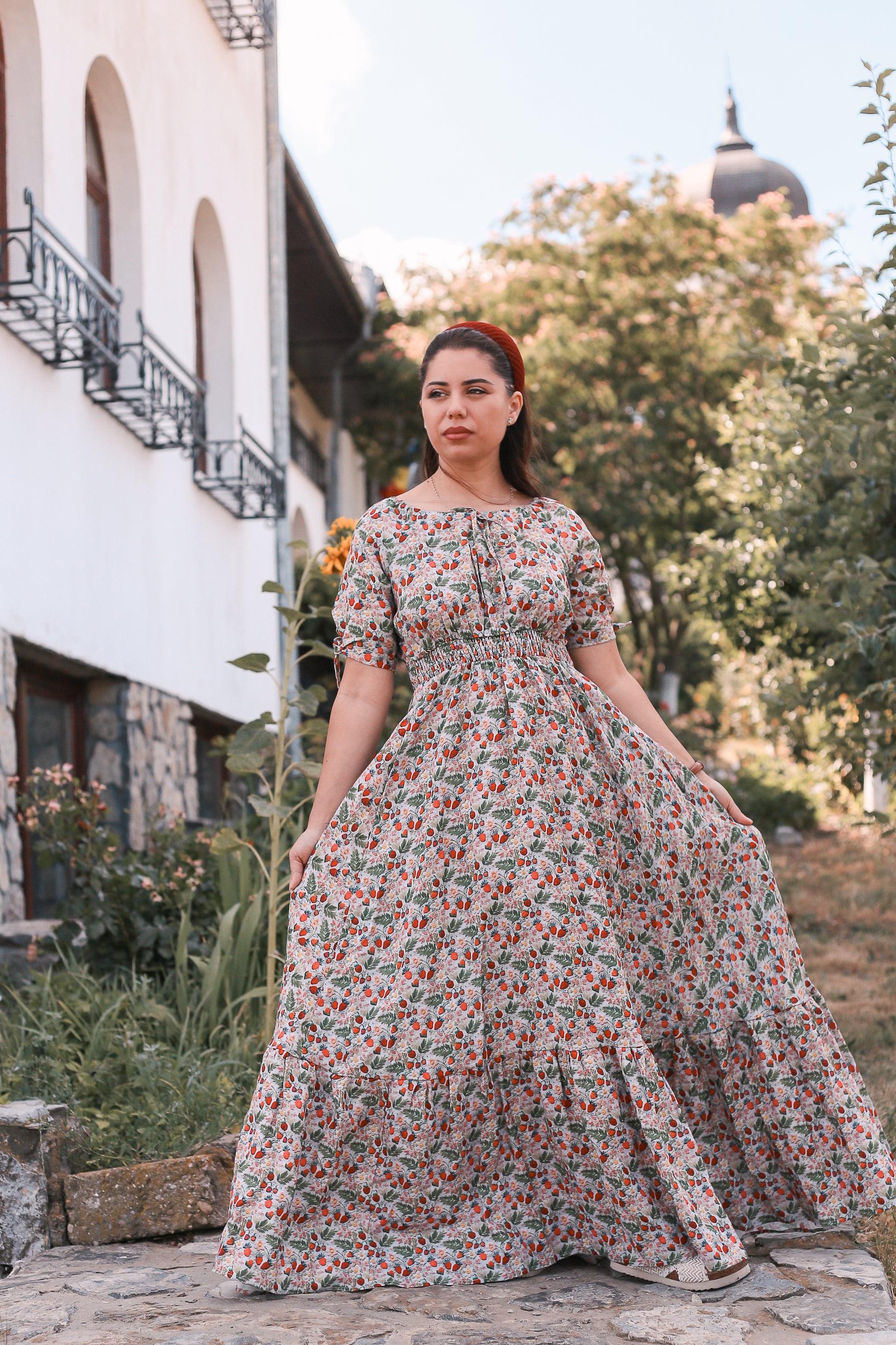 Woman in a floral modest nursing dress standing outdoors with a building and trees in the background