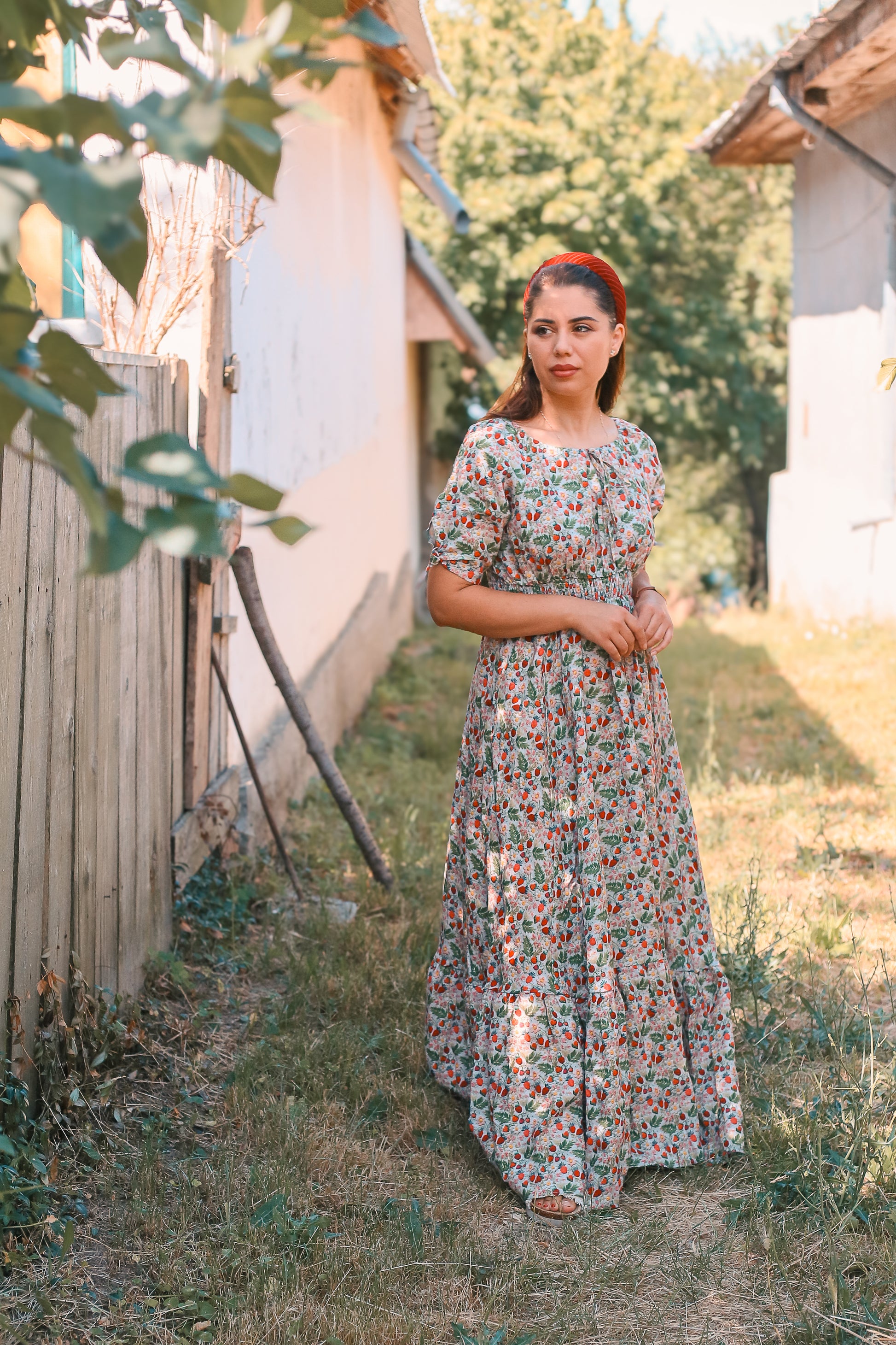 Woman in a floral modest nursing dress standing outdoors near a wooden fence and house.