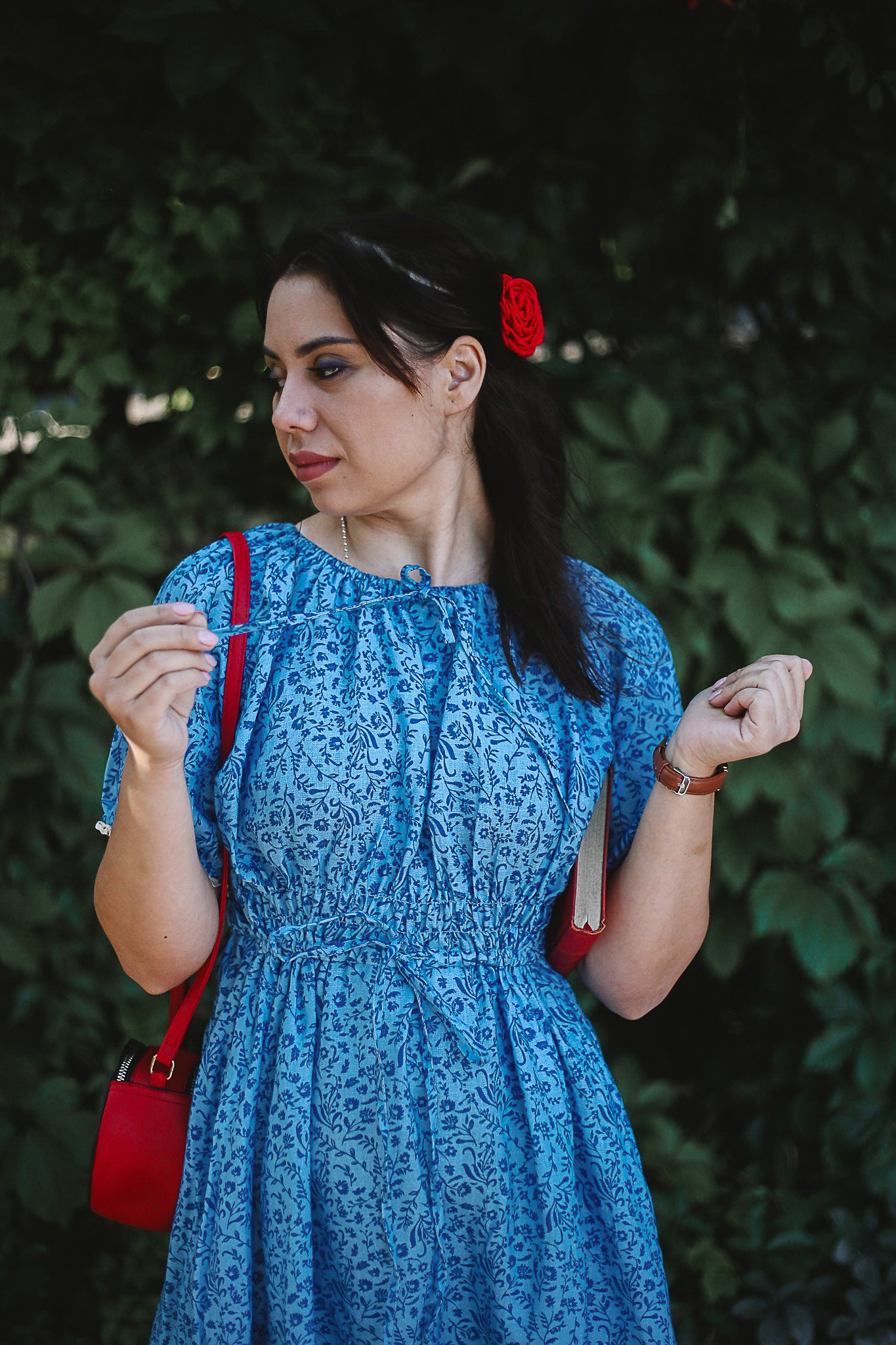 Woman in a blue modest nursing dress with a red bag and hair clip against a green leafy background