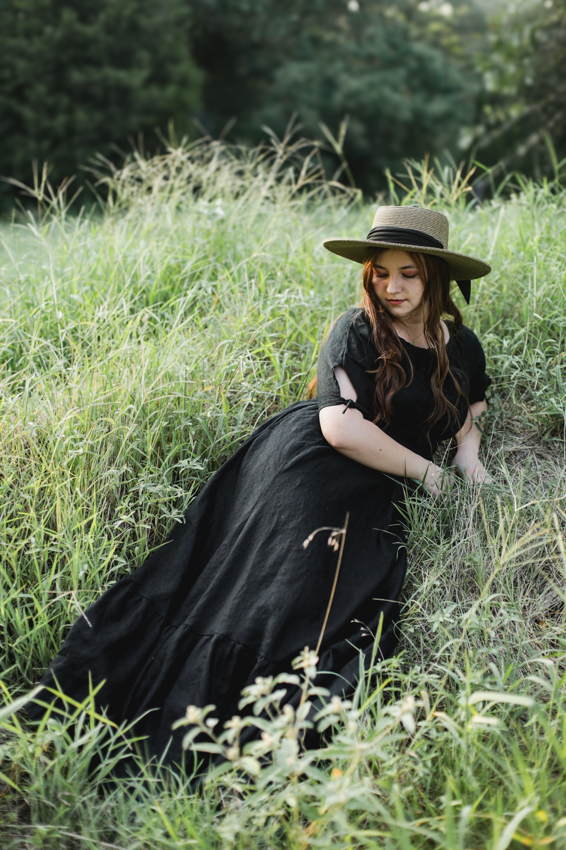 Woman in a black modest nursing dress and hat sitting in a grassy field