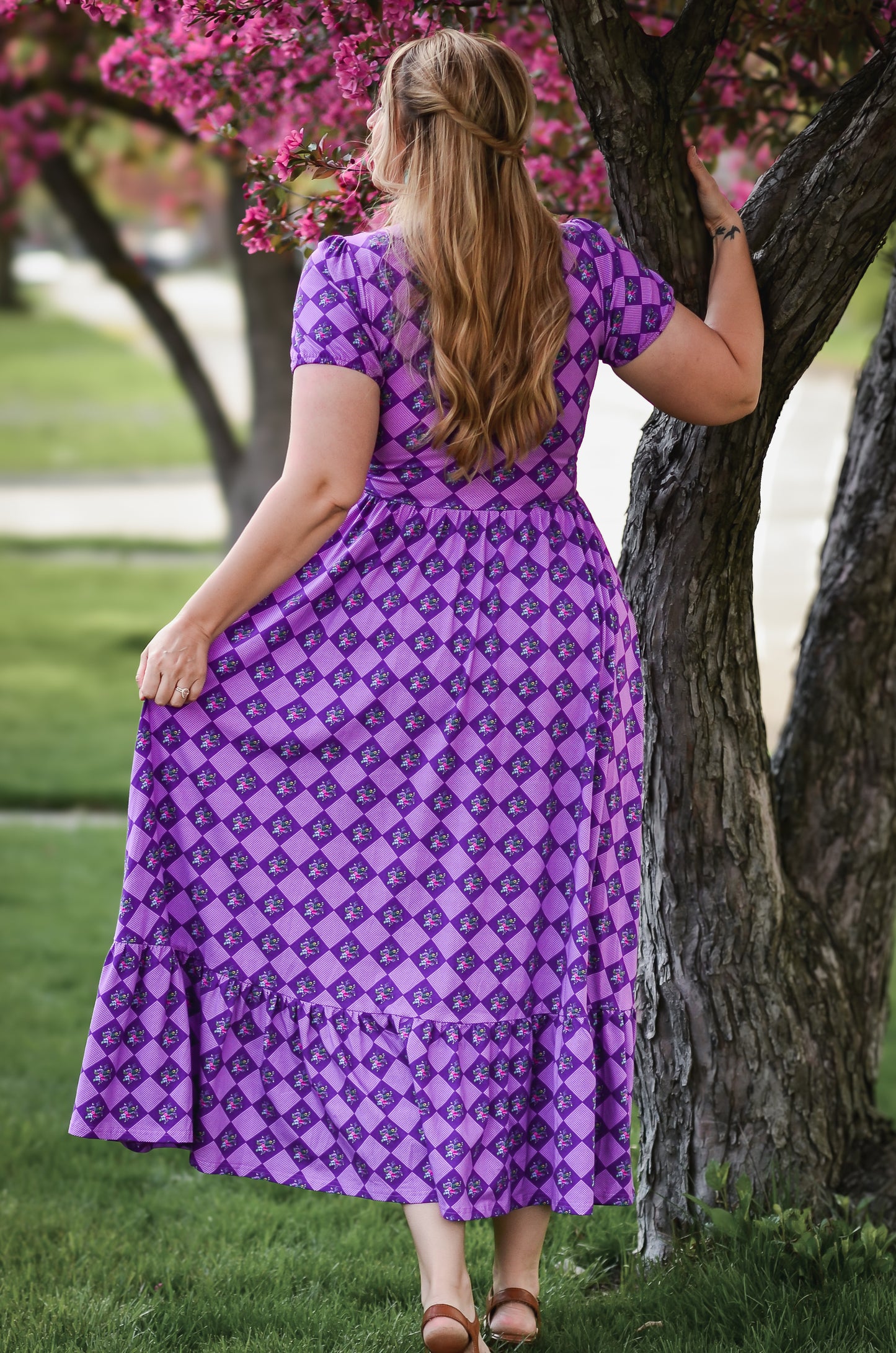 Woman in a purple patterned dress standing by a tree with pink flowers.