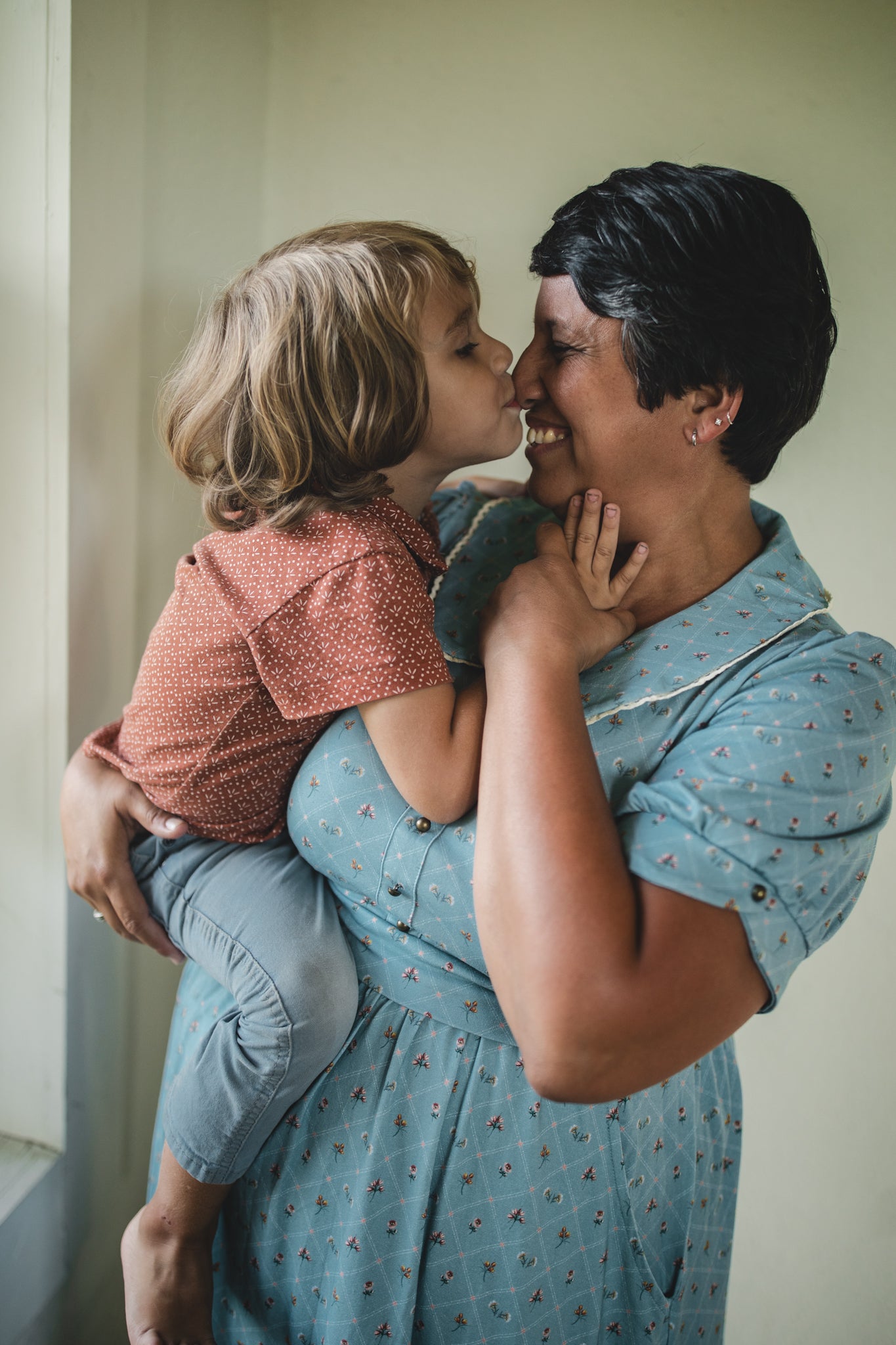 Woman in a blue modest nursing dress holding a child, both smiling and embracing.